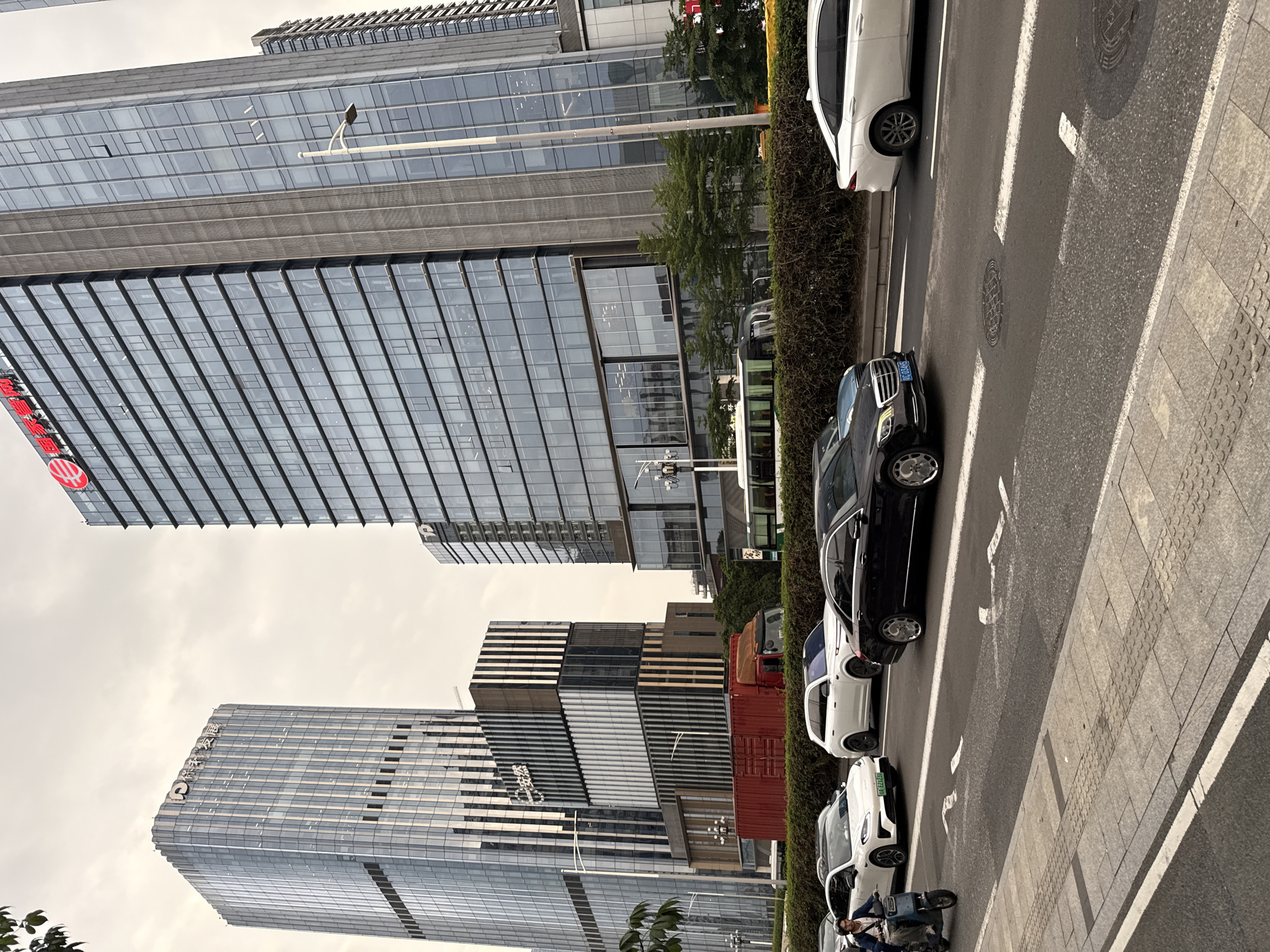 In Tianhe District, China, a street scene unfolds with modern architecture and urban activity under an overcast sky. Dominating the background are several contemporary high-rise buildings with glass facades. The tallest building on the left features a prominent logo and vertical lines. Adjacent to it, a shorter, tiered building displays vertical striped patterns. To the right, another imposing glass-fronted skyscraper displays a red circular logo at its apex.
In the foreground, a multi-lane road carries traffic. A red shipping container is visible behind a low-lying hedge. Several cars are in motion, including a white sedan and a black sedan with chrome wheels. On the left side of the road, a person is riding a blue electric scooter, wearing a blue jacket and dark pants. The road has white lane markings and a pedestrian walkway with textured paving stones is visible on the far left. A white bus is also present on the road, behind the hedge. Green trees are interspersed with the buildings, adding a touch of nature to the urban landscape.