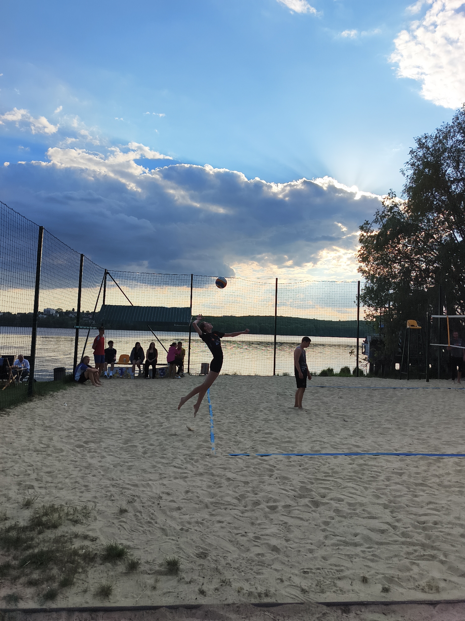 The image depicts a beach volleyball scene at Ternopil Lake in Ternopil, Ukraine. A young woman is in mid-air, hitting a volleyball over the net. A male player stands on the opposite side of the net. In the background, a group of people sit on a bench. A lake and trees are visible behind the volleyball court. The sky is blue with clouds, and the sun is setting. The volleyball court is made of sand. A fence surrounds part of the court.