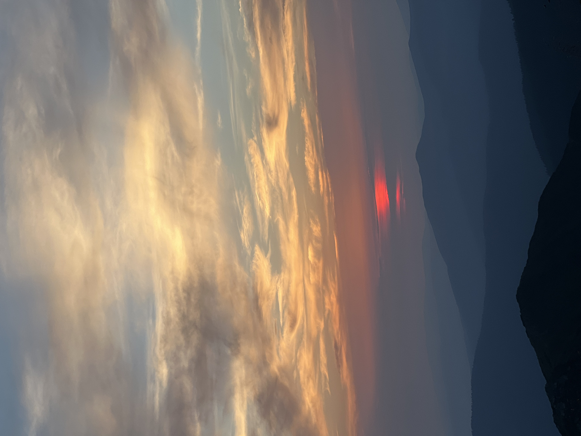 A landscape photograph depicts a hazy mountain range at dusk. The sky is filled with wispy clouds painted in hues of orange, yellow, and blue, indicating the setting sun. The sun itself appears as a vivid red orb peeking through the atmospheric haze. Layered mountains, silhouetted in shades of blue and purple, recede into the distance. The foreground features a dark, sharply defined mountain peak. The overall impression is one of natural beauty and tranquility. The image does not contain any people, animals, or man-made structures, and therefore, cannot be specifically located in Luhy, Ukraine, or any other urban area.