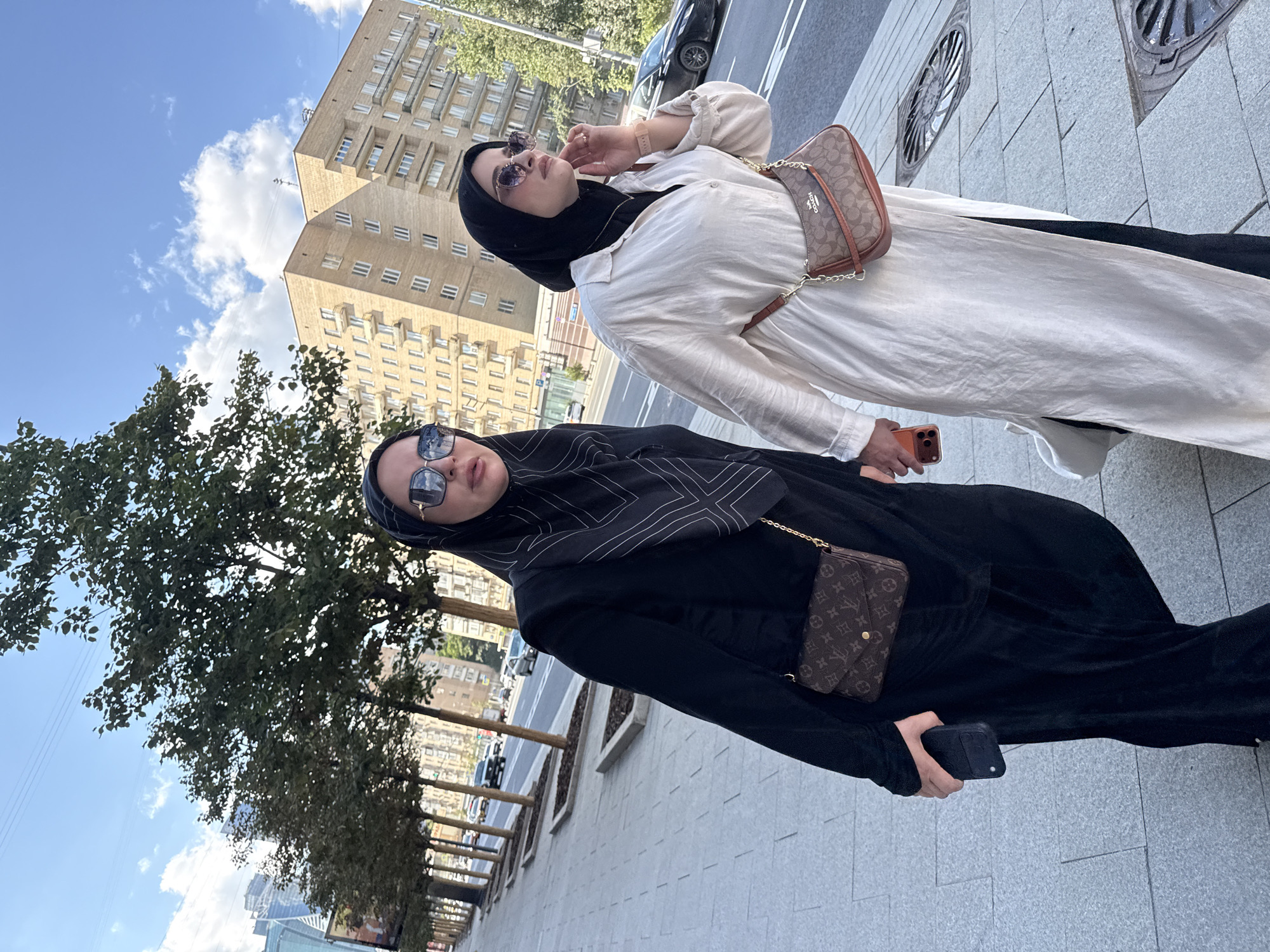 Two women, dressed in traditional Islamic attire and sunglasses, walk side-by-side on a city sidewalk. The woman on the left wears a black abaya and hijab with a patterned scarf, carrying a black phone and a brown Louis Vuitton purse. The woman on the right wears a white abaya over black clothing, carrying a Coach purse and holding an orange phone. They are walking on a paved sidewalk with trees and a street with cars in the background. Behind them, a light-colored, multi-story building with many windows dominates the cityscape. The sky is blue with scattered clouds. The scene appears to be a sunny day in Moscow, Russia, likely in a residential or commercial area with a tree-lined street.