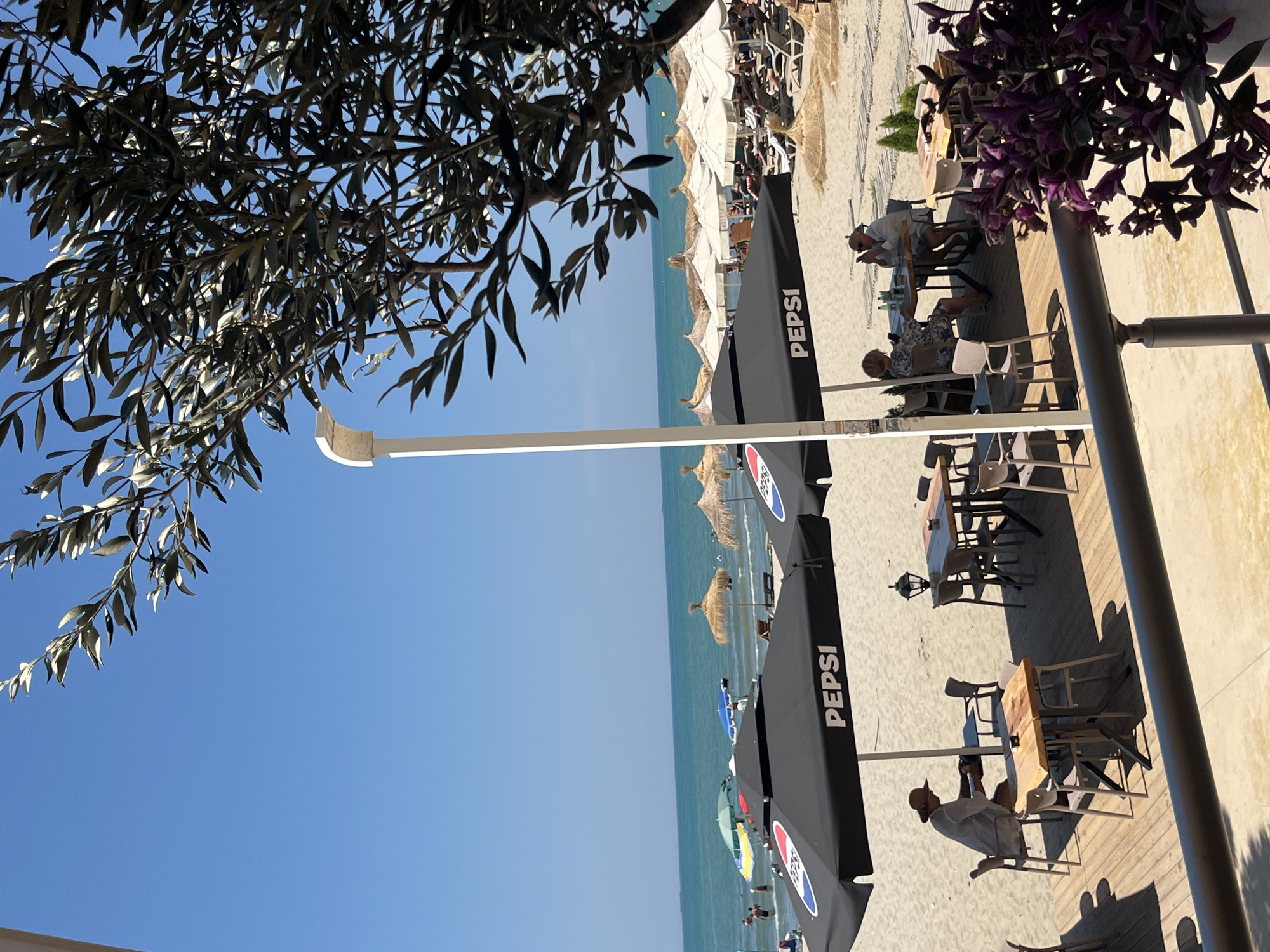 This image depicts a beach scene in Bashkia Durrës, Albania. In the foreground, there is a wooden deck with tables and chairs, shaded by large black umbrellas with "PEPSI" branding. Two people are seated at one of the tables, looking at their phones. In the middle ground, more beach umbrellas, some made of straw and others white, are set up on the sand. People are visible in the shallow water of the turquoise sea, and lounge chairs are arranged on the beach. The background shows a clear blue sky and the vast expanse of the sea. A tree with dark leaves partially obstructs the upper right portion of the image, casting shadows.