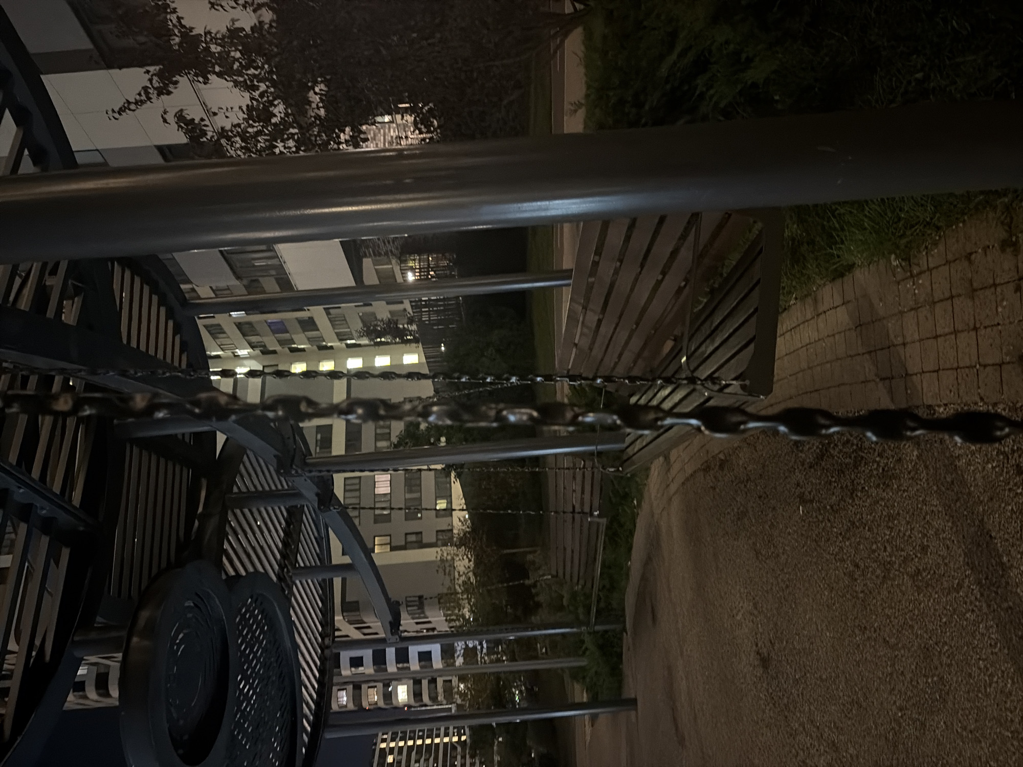 The image captures a dimly lit outdoor area, possibly a playground or park, at night. A chain hangs prominently in the foreground, suggesting the presence of swings. In the background, modern apartment buildings with illuminated windows are visible, indicating an urban setting. The architecture of the buildings and the overall ambiance suggest a location within a planned residential development in Moscow, Russia. The ground appears to be a mix of gravel and paving stones. Lush green bushes and trees are also present, adding to the natural elements of the environment. The scene is devoid of any people, and no overt actions or interactions are taking place. The lighting is artificial and creates long shadows, contributing to a quiet and somewhat secluded atmosphere.