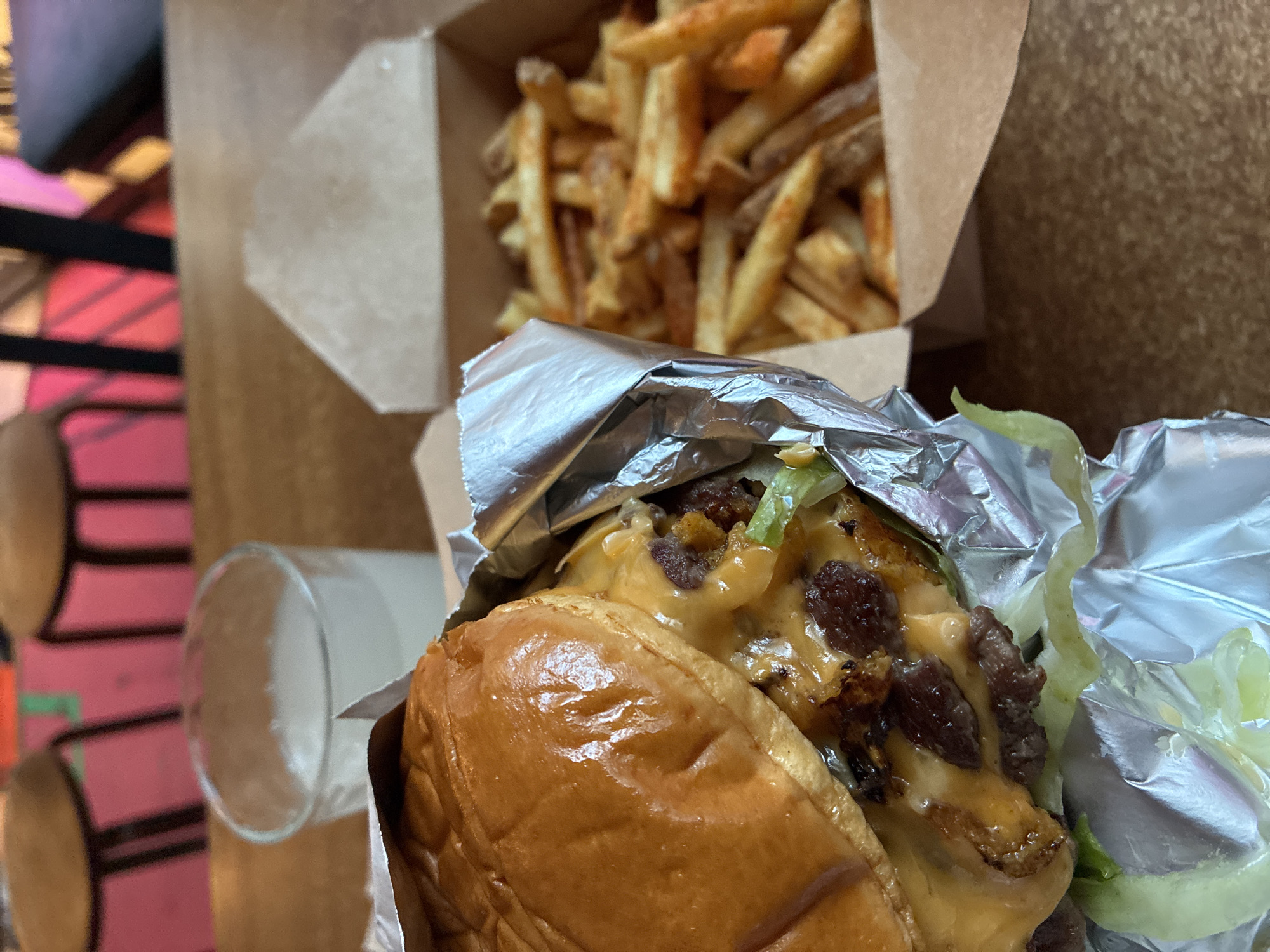 This image presents a close-up, eye-level view of a meal placed on a wooden table within a restaurant environment. In the foreground, positioned to the left, is a burger partially unwrapped from its encompassing aluminum foil. The burger features a glossy, golden-brown brioche bun, two smash-style beef patties covered with melted yellow cheese, and visible green lettuce leaves beneath. To the right of the burger, an open brown cardboard box contains a quantity of seasoned french fries. A clear glass, partially filled with an opaque white liquid (likely milk or a milkshake), is situated centrally behind the burger. The background is out of focus, revealing the tops of wooden chairs with pink upholstery and sections of a wall with abstract patterns in vibrant pink, green, and dark colors. The distinctive culinary presentation, including the burger style and packaging, combined with the discernible interior design elements, identifies the location as a Goiko Grill restaurant, situated in the city of Barcelona, Spain.