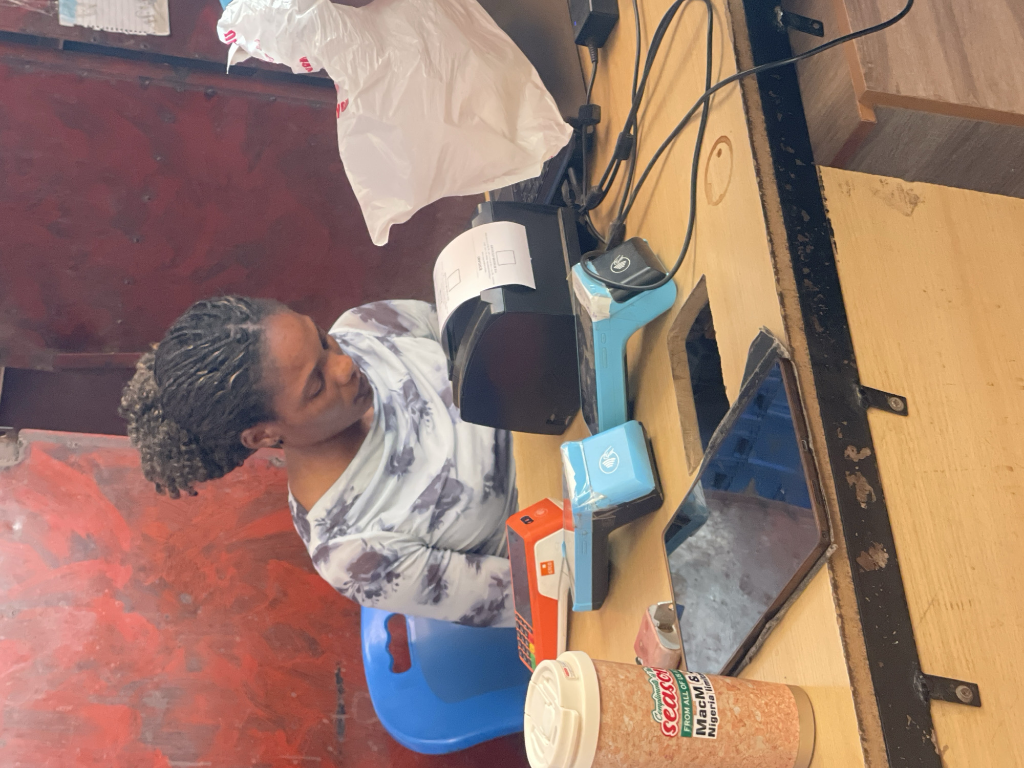 The image shows a woman with braided hair sitting on a blue plastic chair in Gura Top, Nigeria. She's next to a countertop holding a black printer, a blue credit card reader, a small orange box, a tablet, and a cylindrical container labeled "Seaco." A white plastic bag hangs from above the printer. The counter and wall behind it are light brown with dark trim. The area adjacent to the chair and the wall are a red and grey mottled color. Electrical cords are visible near the printer.