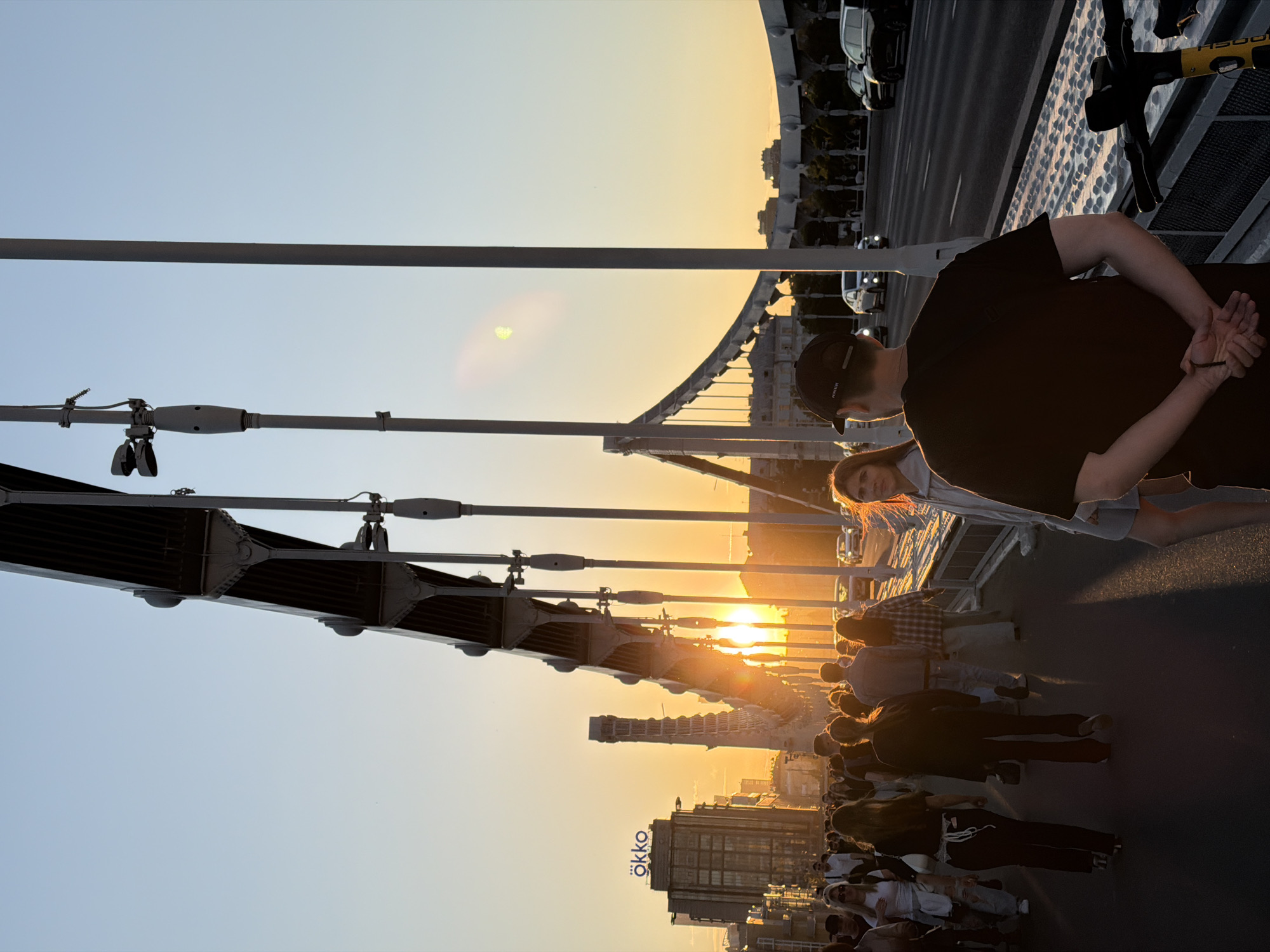 A crowded pedestrian bridge in Moscow, Russia, is seen at sunset. The bridge structure features prominent metal supports and overhead wiring. A man in a dark t-shirt and cap is standing with his hands behind his back near a bicycle parked at the edge of the bridge. A woman stands next to him, visible just below his shoulder. The sun is setting behind a cityscape, casting a golden light on the buildings. Below the horizon, there is a modern building with a sign that reads "ökko." A line of cars is visible on the roadway parallel to the bridge. Many pedestrians are walking on the bridge.