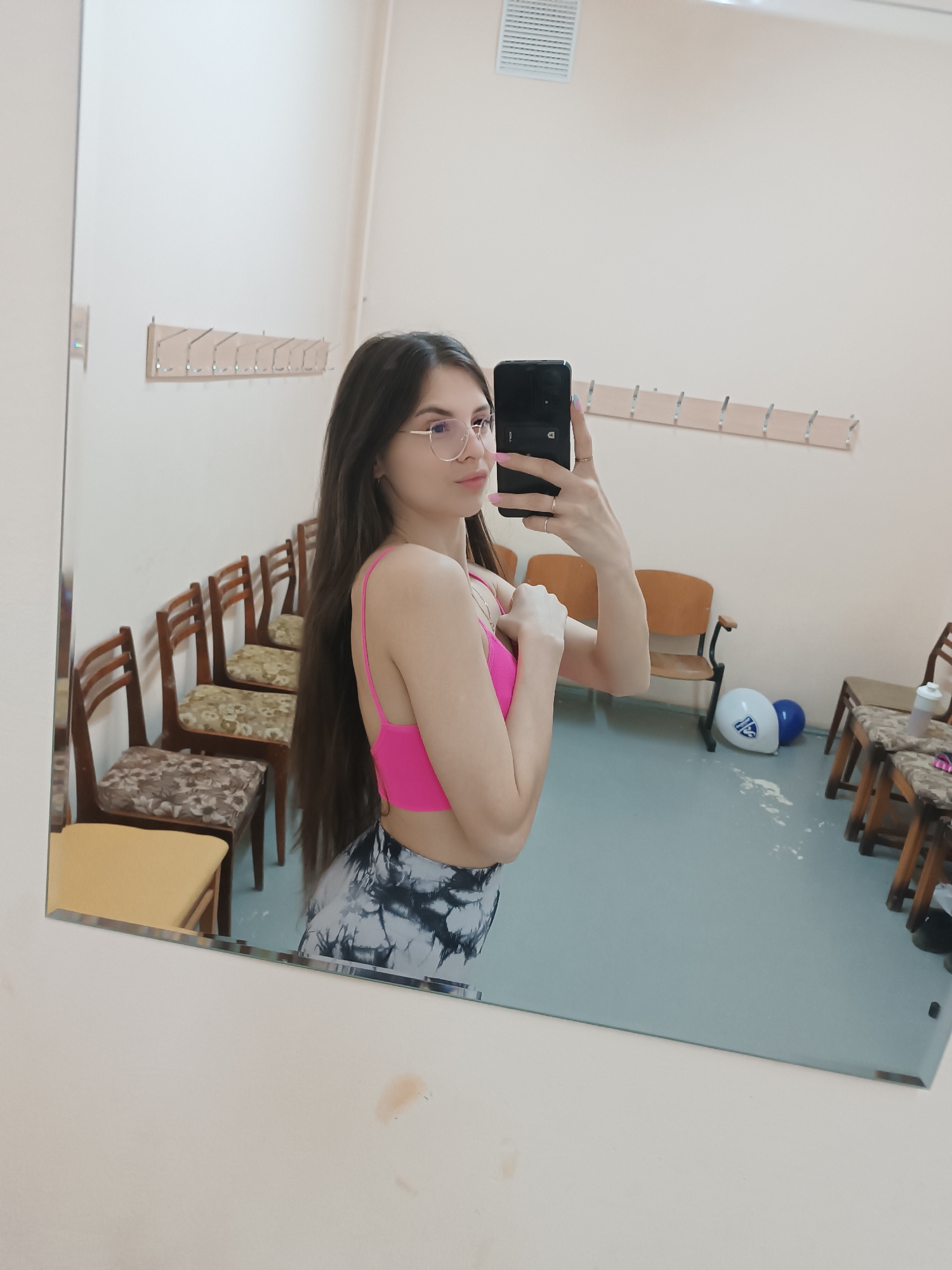 A mirror reflects a young woman in a changing room. She is wearing a pink sports bra and patterned leggings, holding a phone to take a selfie. Her hair is long and brown, and she wears glasses. The changing room contains rows of chairs with patterned cushions, hooks on the wall, and balloons on the floor.
