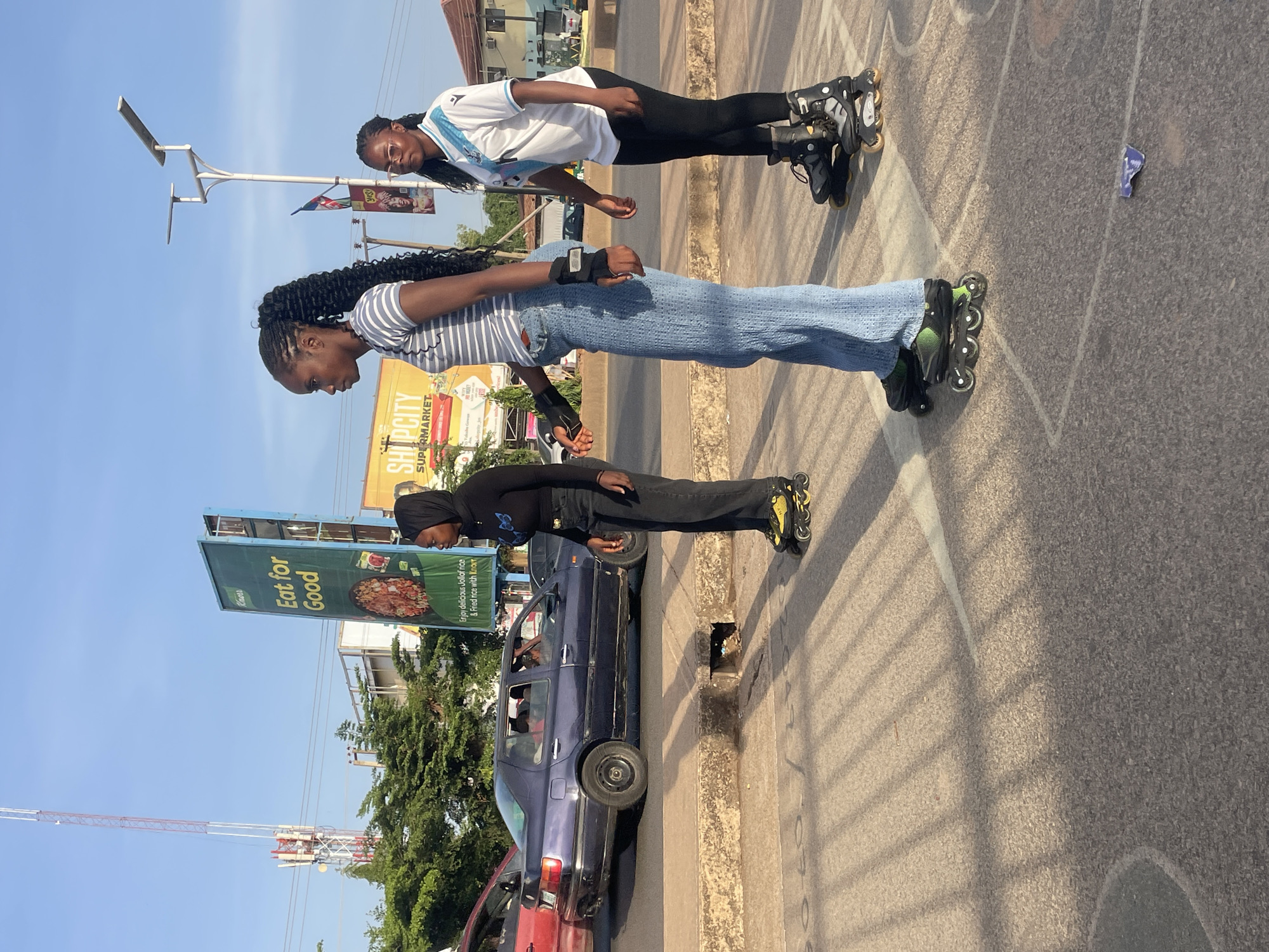 Three individuals are roller skating on a city street near parked cars and billboards in Jos, Nigeria.
The first person is wearing a white jersey with blue accents, paired with black leggings and black roller skates. They are positioned towards the top of the frame.
The second person is wearing a striped top with light blue jeans, wrist protection, and black roller skates. They are in the middle of the frame.
The third person, located near the bottom of the frame, is wearing a black top with a hijab and black pants. They are also wearing roller skates, with yellow accents. They are standing near a parked blue car.
In the background, there are billboards advertising food. One is a Knorr billboard with the slogan "Eat for Good". Another billboard is visible promoting "SHOPCITY SUPERMARKET". There are overhead lines and a tower extending toward the sky. Street lighting is also present. The day appears to be sunny with clear skies.