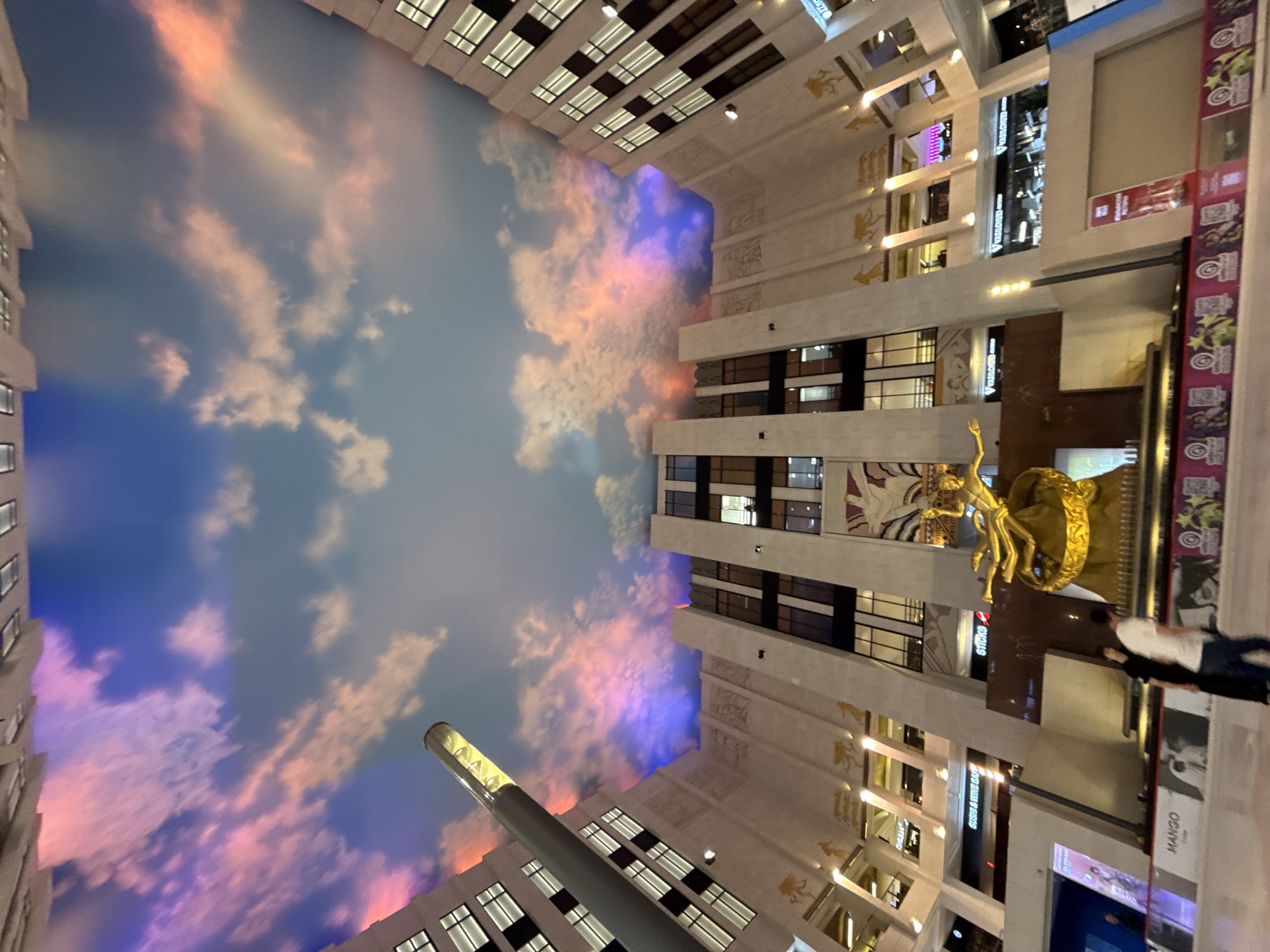 This image, taken in Khimki, Russia, shows a wide-angle view of an interior atrium with a painted sky ceiling. The ceiling depicts a sunset with hues of blue, pink, and orange clouds, creating an illusion of an open sky.
The atrium features multi-story architecture with angular, white facades and numerous windows. Architectural elements include decorative relief carvings on the facades and gold-colored figures or symbols. A tall, cylindrical structure with a light fixture extends upwards from the atrium floor towards the ceiling.
In the center of the atrium, a prominent golden statue is visible, depicting a figure in a dynamic pose, possibly mythological. In front of the statue, two individuals are walking away from the camera. One person is wearing a white t-shirt and dark pants, while the other is in dark clothing.
The lower sections of the atrium show various storefronts with illuminated signs, including "MANGO," "SUSHI & WINE BAR," and "STICKS." A decorative border with colorful graphics and images runs along the base of the storefronts. The overall scene suggests a shopping mall or a large indoor public space with an elaborate design intended to evoke an outdoor atmosphere.