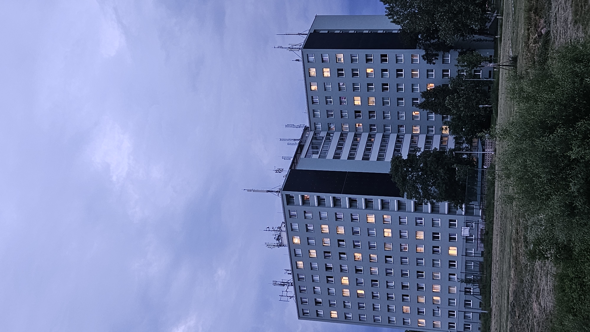 The image shows a tall, light blue apartment building with numerous lit windows, indicating occupancy. The building has a dark blue vertical section and is topped with several antennas. In front of the building, there is a green lawn with some trees and a basketball court with a fence. The sky is overcast with dark, moody clouds. The building is located in the Sekčov housing estate in Prešov, Slovakia.