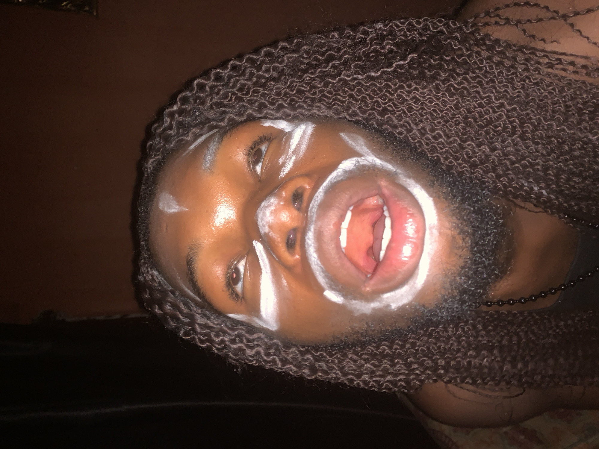 This close-up, angled photograph, potentially taken in Onitsha, Nigeria, captures a person with dark skin and long, tightly curled, dark hair or braids. The individual is positioned looking upwards and slightly to the left from the viewer's perspective, with their mouth wide open, revealing the tongue and teeth. This suggests an act of vocal expression such as yelling, singing, or speaking intensely.
The person's face is adorned with distinct white paint in a decorative pattern. Streaks are visible on the forehead, below both eyes, around the nose, and a thick, circular outline frames the open mouth. Fine facial hair is apparent around the chin and jawline, suggesting the subject is an adult male. A black beaded necklace can be seen partially on the right side of the frame, resting against the neck.
The background is dark and indistinct, implying a low-light indoor setting or nighttime. The lighting highlights the person's skin, particularly the forehead and cheeks, and makes the white face paint stand out. The image conveys a sense of cultural performance, ritual, or artistic expression. No text is visible within the media file.