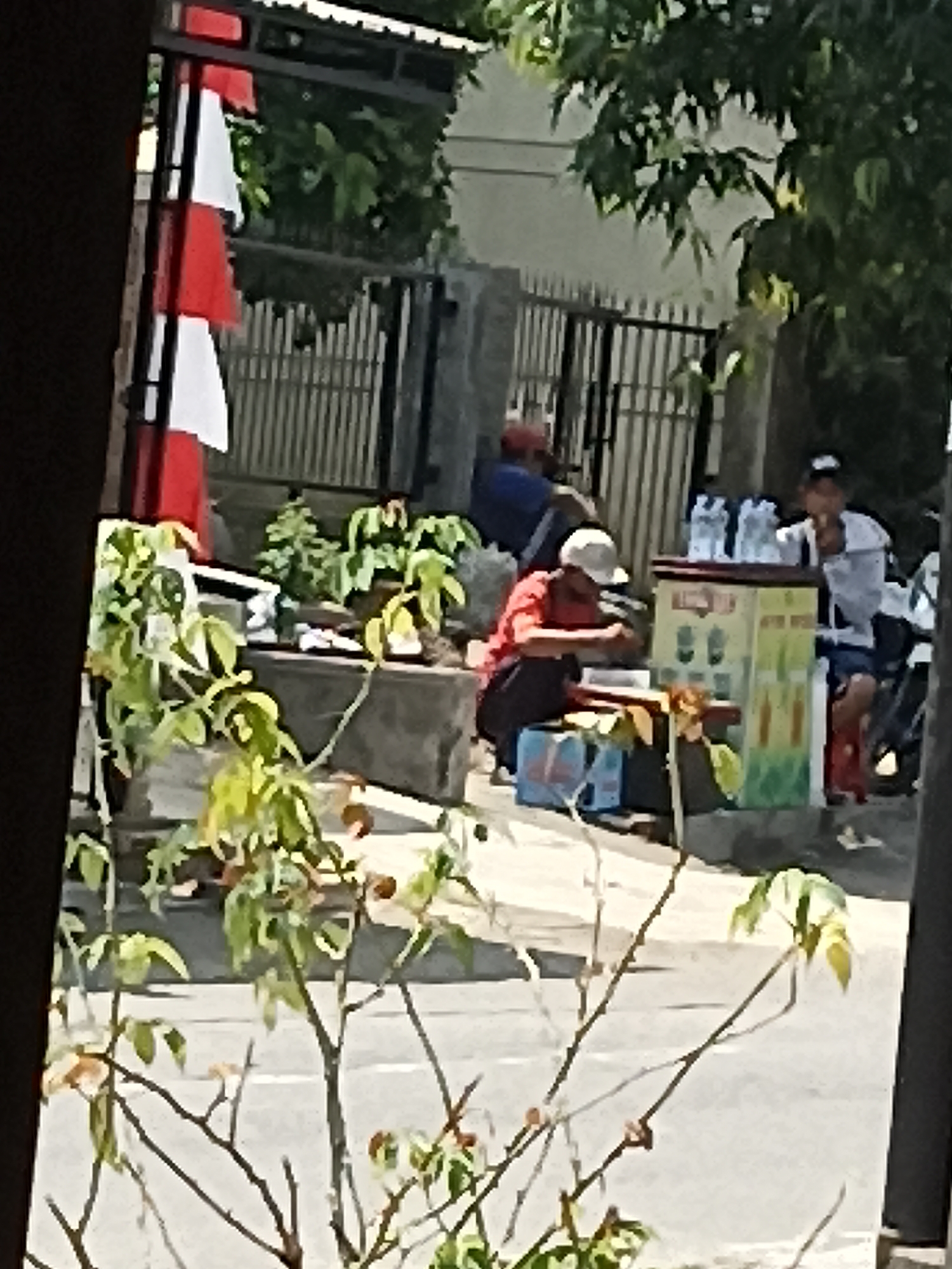 In Pulisen, Indonesia, an outdoor scene unfolds with several individuals near a roadside stall. A person in a red shirt and white cap is crouched, seemingly preparing something. To their right, another individual in a white shirt and dark shorts sits beside a colorful, multi-paneled cart that displays beverages. Several bottles of water are visible on top of the cart. Behind them, a motorcycle is parked. Further back, a metal fence encloses an area with greenery. A flag with alternating red and white stripes is mounted on a pole near the left edge of the frame. The environment suggests a public space, possibly a street or sidewalk, with trees and foliage providing some shade.