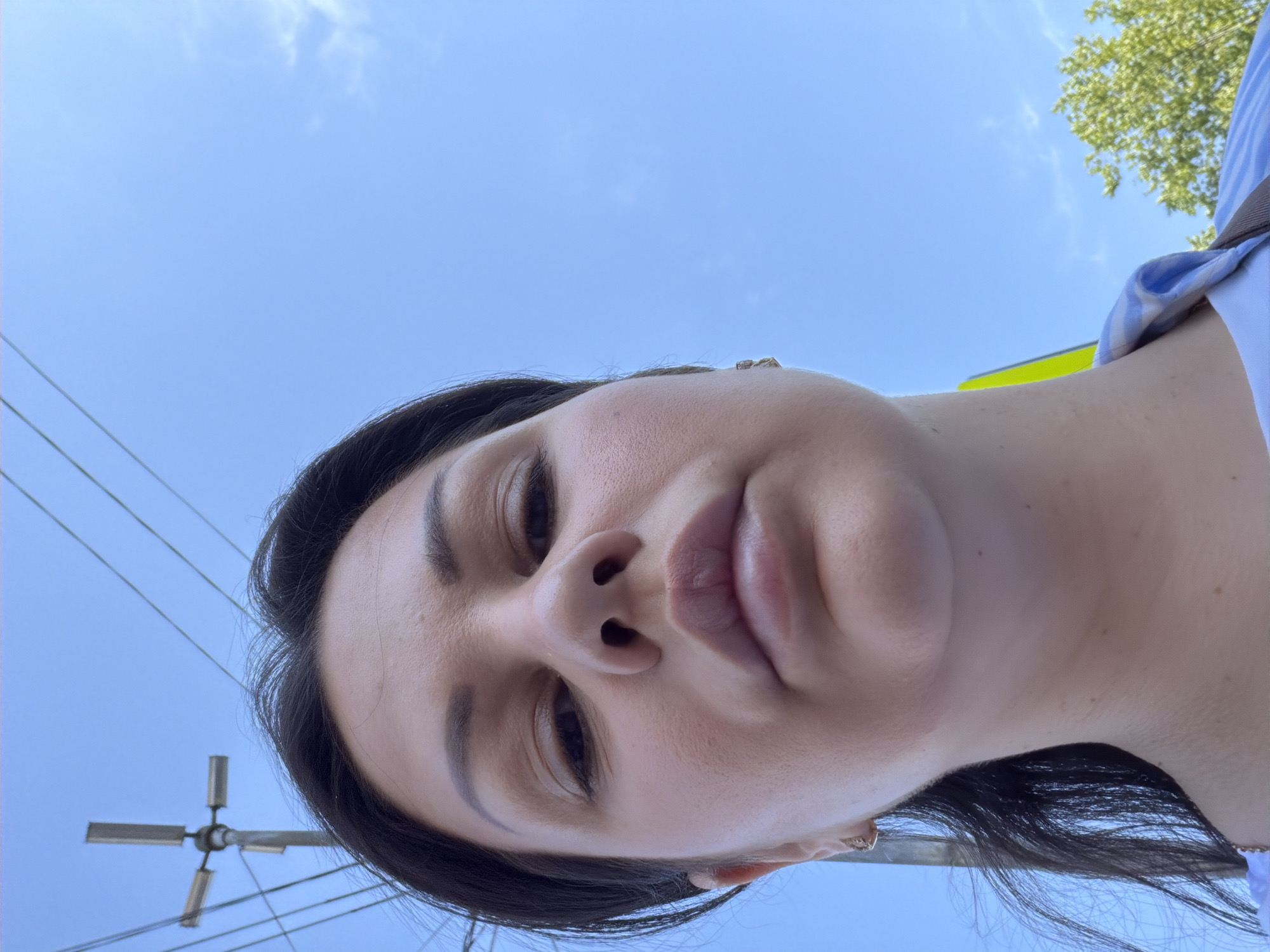 A low-angle selfie depicts a woman outdoors under a clear blue sky with minimal cloud cover. The woman has dark hair, pronounced eyebrows, closed or downcast eyes, and full lips. She wears small earrings in both ears, and a light blue and white striped collar with a dark strap is visible on her right side. In the background, to the upper left, a utility pole with a multi-directional light fixture stands, surrounded by multiple black overhead wires. Green tree foliage is visible in the lower right background. No specific landmarks or features within the image visually confirm the location as Sochi, Russia.
