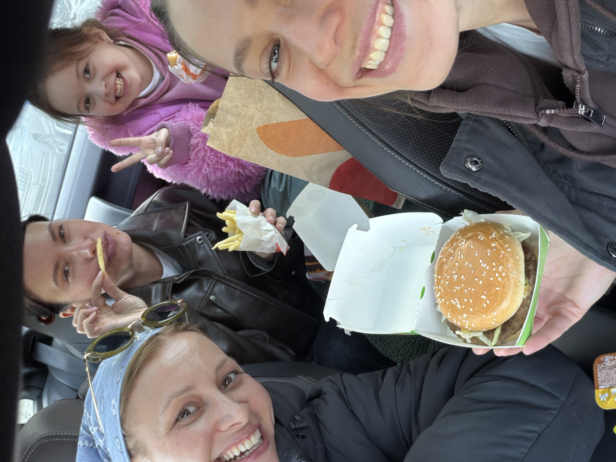 This image depicts four females—three adults and one child—inside a vehicle in the city of Khimki, Russia. The scene captures a moment of fast-food consumption. A woman in the immediate foreground, positioned right, holds a burger in a white and green carton, her smiling face partially visible as she takes a selfie. Seated to her left, another adult female, wearing a blue bandana and sunglasses on her head, smiles towards the camera. Further back in the vehicle, an adult female in a dark leather jacket holds a single french fry to her lips while also holding a paper bag containing more fries. Beside her, a young girl in a pink fluffy jacket smiles and displays a 'peace' sign with her right hand. A Burger King paper bag is visible between the two occupants in the deeper part of the frame. The vehicle's interior features dark upholstery. An indistinct exterior view through the rear window suggests an outdoor, possibly winter, environment. A small yellow condiment packet rests on the lower left.