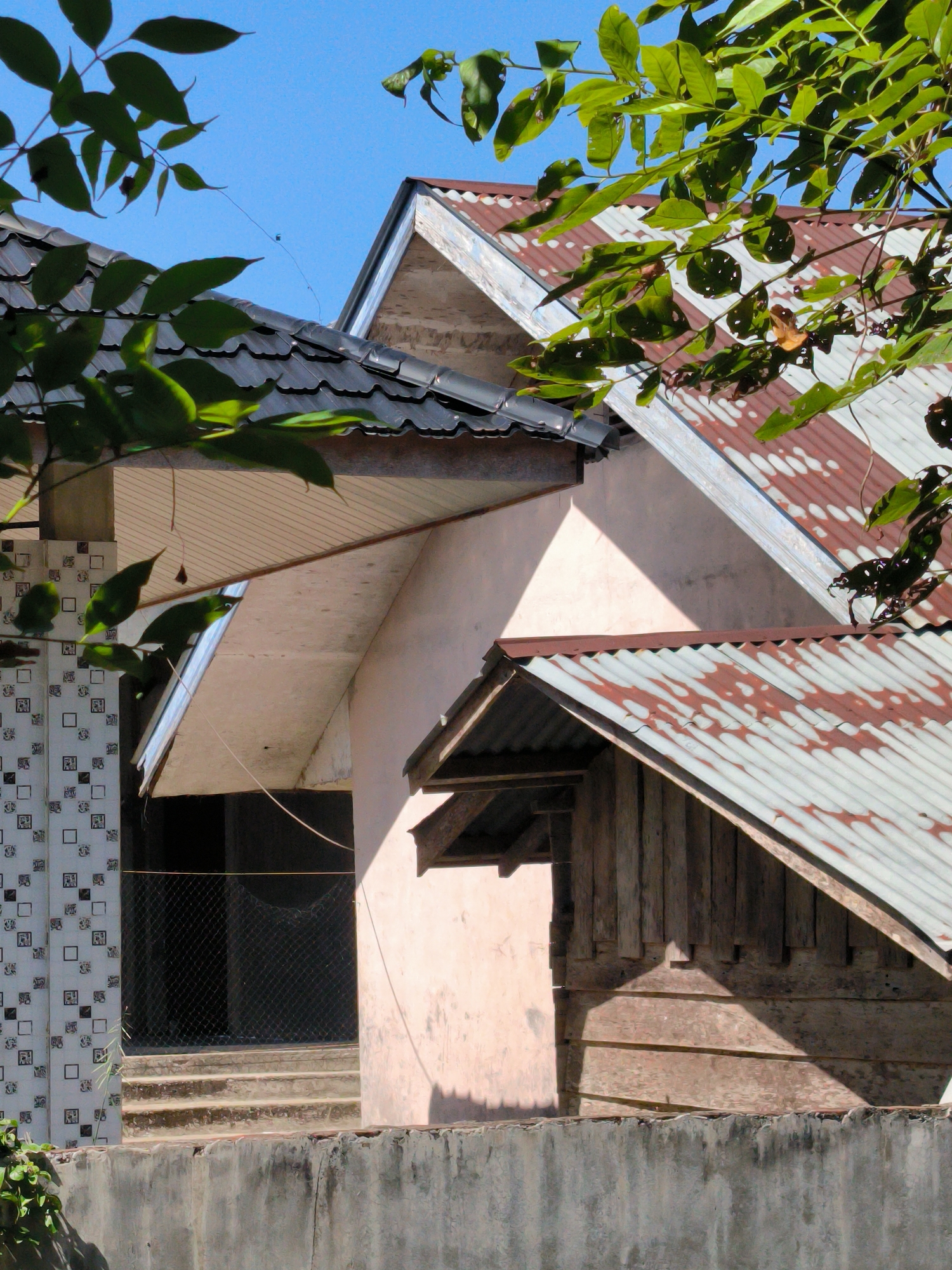 A sunlit exterior view shows a collection of buildings in Merbopuntong, Indonesia. The structures display a mix of architectural styles and materials, including corrugated metal roofs with rust patterns, tiled roofing, concrete walls, and wooden siding. A wall with tiled accents is also visible. Lush green foliage frames the scene, partially obscuring the buildings and contrasting with the blue sky. The scene is still, with no evident human activity.