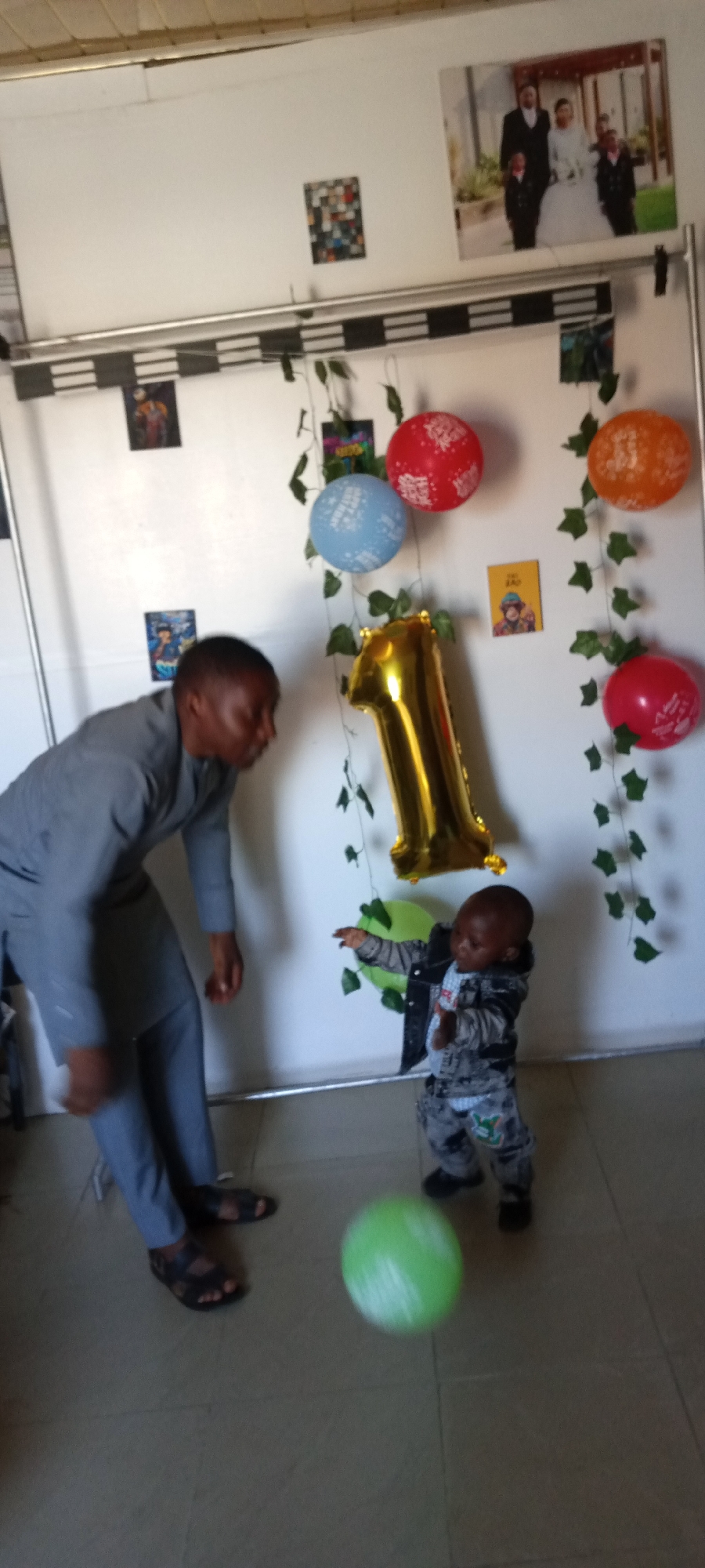 A photo shows a man and a young child in a room decorated for a first birthday. The man is wearing a gray suit and sandals, bending towards the child. The child, wearing a patterned outfit and a jacket, is standing and reaching towards a green balloon that is mid-air. The backdrop features a white wall adorned with balloons (red, blue, orange), artificial ivy garlands, small photographs, and a large gold balloon shaped like the number "1". At the top of the wall, above the decorations, are more pictures, including a wedding photo. The floor is tiled. Based on context, the location is likely Jos, Nigeria.