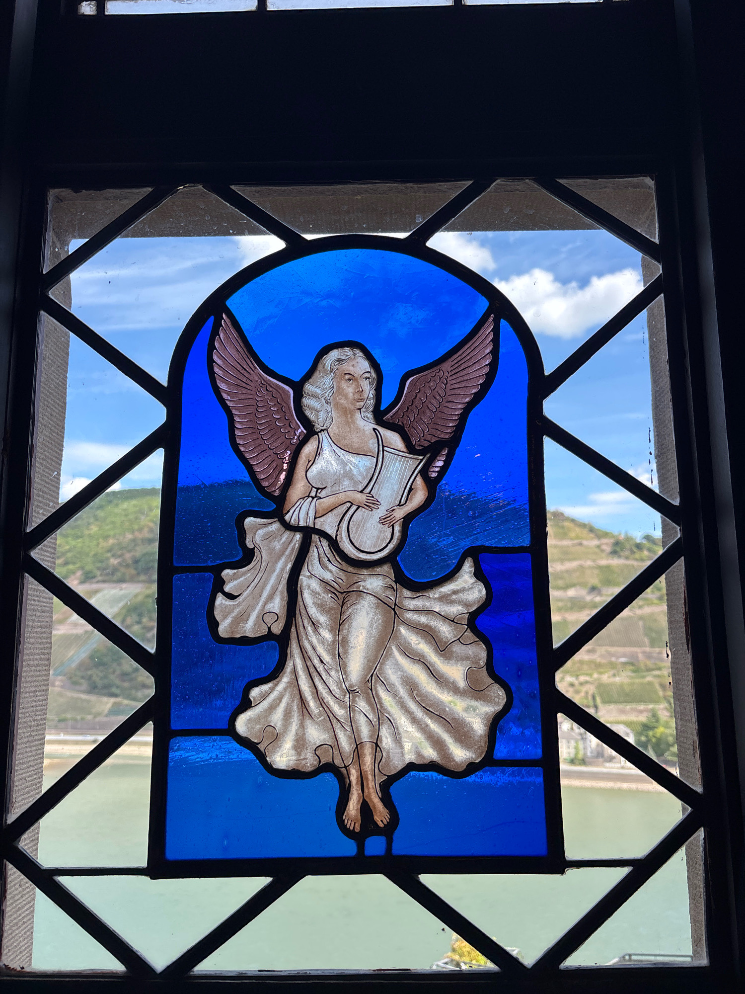 The image shows a stained-glass window depicting an angel playing a lyre. The angel has blonde, curly hair, large, feathered wings, and is wearing a flowing white robe. The background of the stained glass is a vibrant blue. The window itself is framed by dark leaded glass panes, forming a diamond pattern.
Through the stained glass, a scenic view of Assmannshausen, Germany, is visible. This includes the Rhine River, a hillside covered with vineyards, and a building nestled within the landscape. The sky above is blue with scattered white clouds. The window appears to be set within a stone or dark wooden frame.