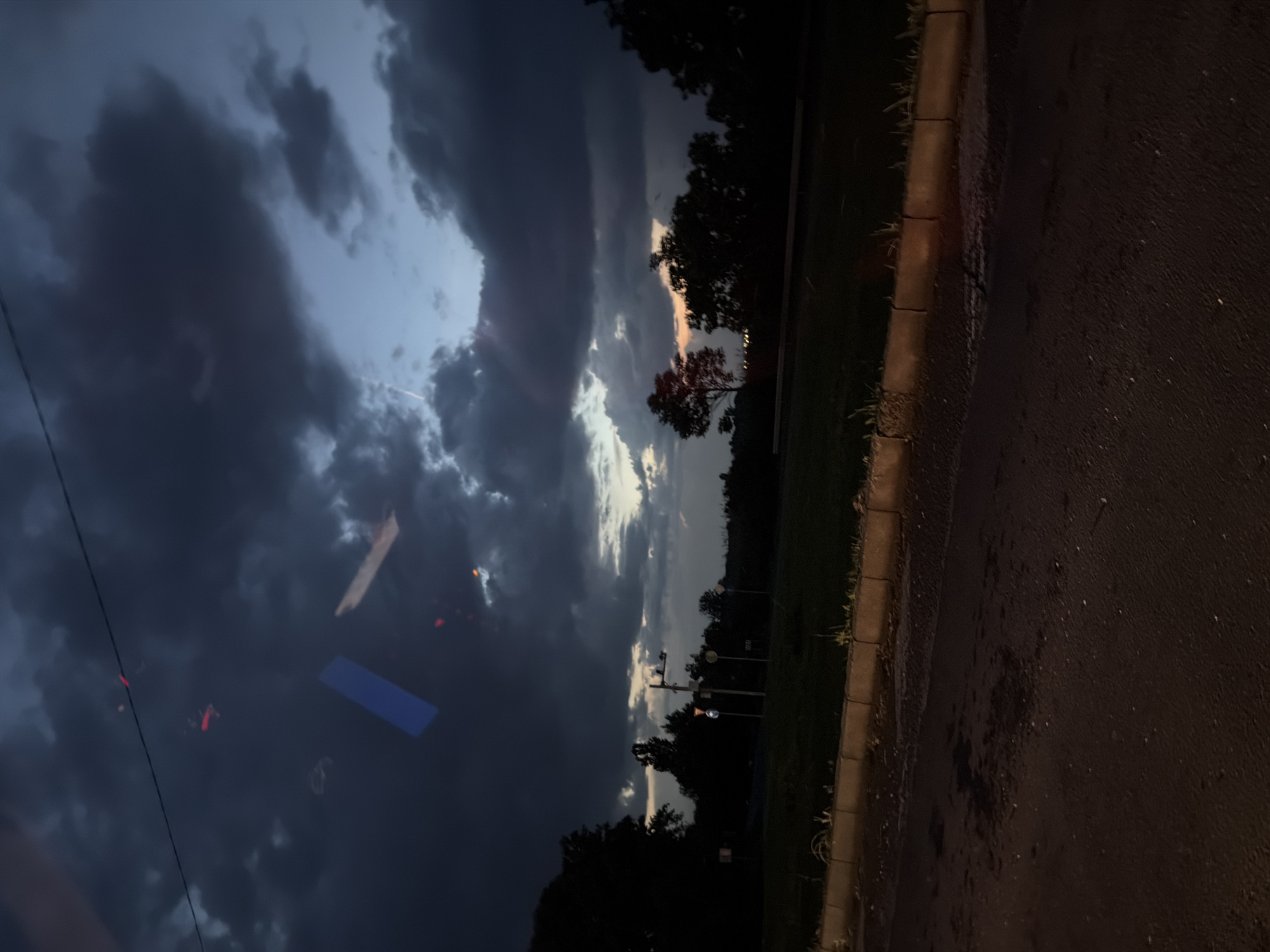 The image captures a view of a dark, stormy sky in Burgas, Bulgaria. Cumulonimbus clouds dominate the sky, obscuring much of the daylight. A section of the horizon reveals a sliver of lighter clouds with a hint of orange, indicating either sunrise or sunset. In the foreground, there are silhouettes of trees and vegetation. A paved area, possibly a street or parking lot, is visible on the right side of the image. The overall impression is one of impending rain and atmospheric drama. The image is rotated counter-clockwise. There appear to be lights or reflections on the glass through which the picture was taken.