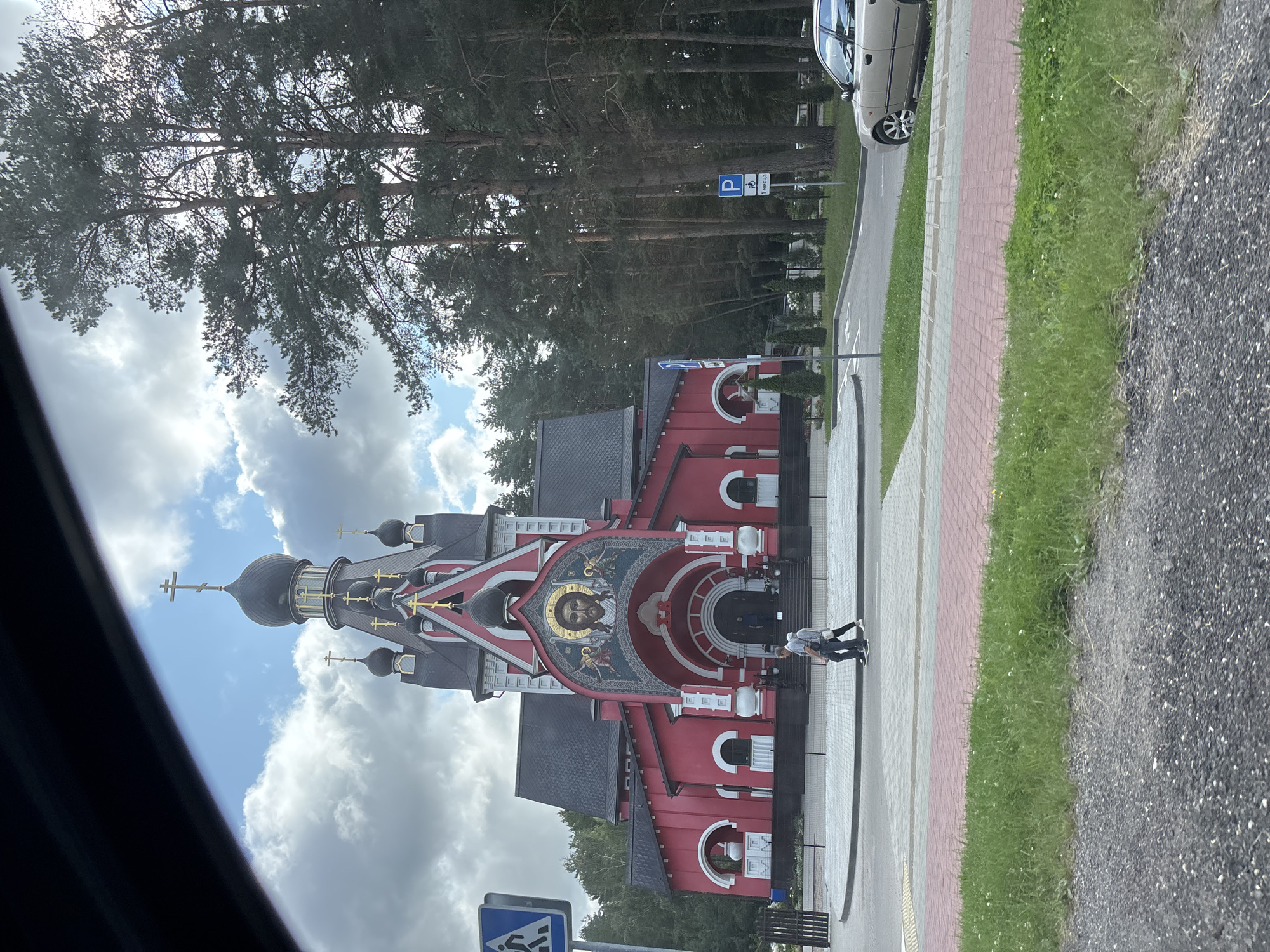 The image depicts a bright red church with black domes and gold crosses, set against a backdrop of tall pine trees and a cloudy sky. The church features a prominent mosaic above its arched entrance, depicting Jesus Christ with accompanying angels. In front of the church, a paved walkway is visible with two individuals walking across it. To the left, a blue pedestrian crossing sign is present, and to the right, a parking sign for disabled persons is displayed. A light-colored sedan is parked on the right side of the frame, near the road. The overall scene appears to be an exterior view of a religious institution in a semi-urban or suburban setting.