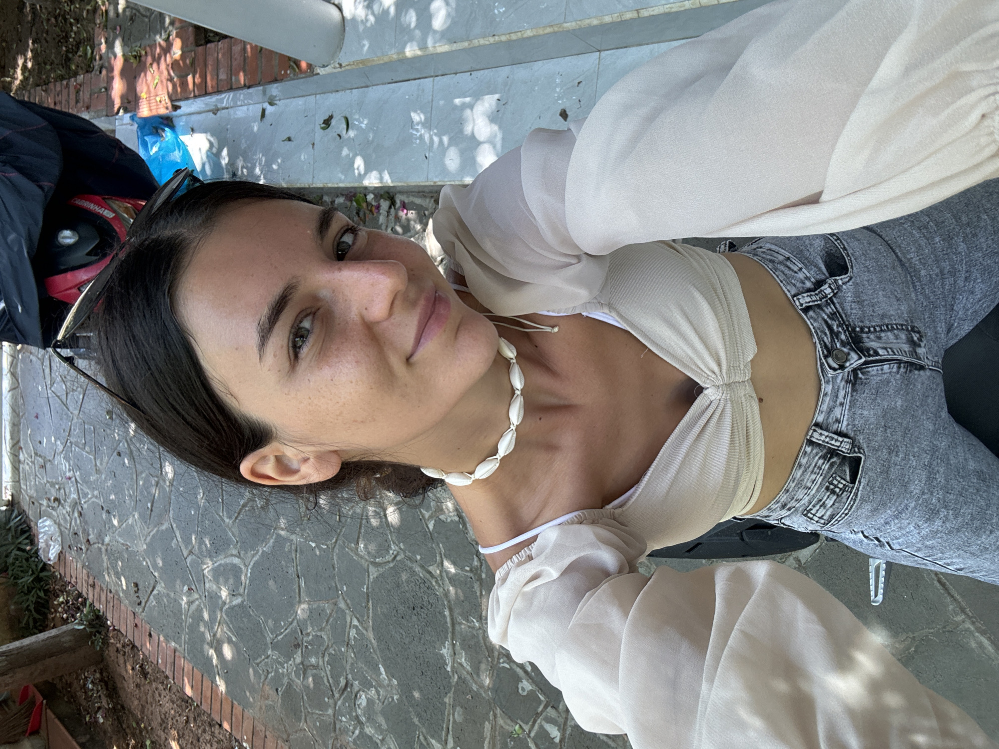 A young woman with dark hair and freckles is taking a selfie while sitting on a motorcycle in Mũi Né, Vietnam. She is wearing a cream-colored crop top with puffed sleeves and a shell necklace. Her lower half is clad in grey jeans. The background shows a paved walkway with shadows cast by trees and sunlight. A red scooter is partially visible behind her. The environment suggests a warm, sunny day in a residential or outdoor area.