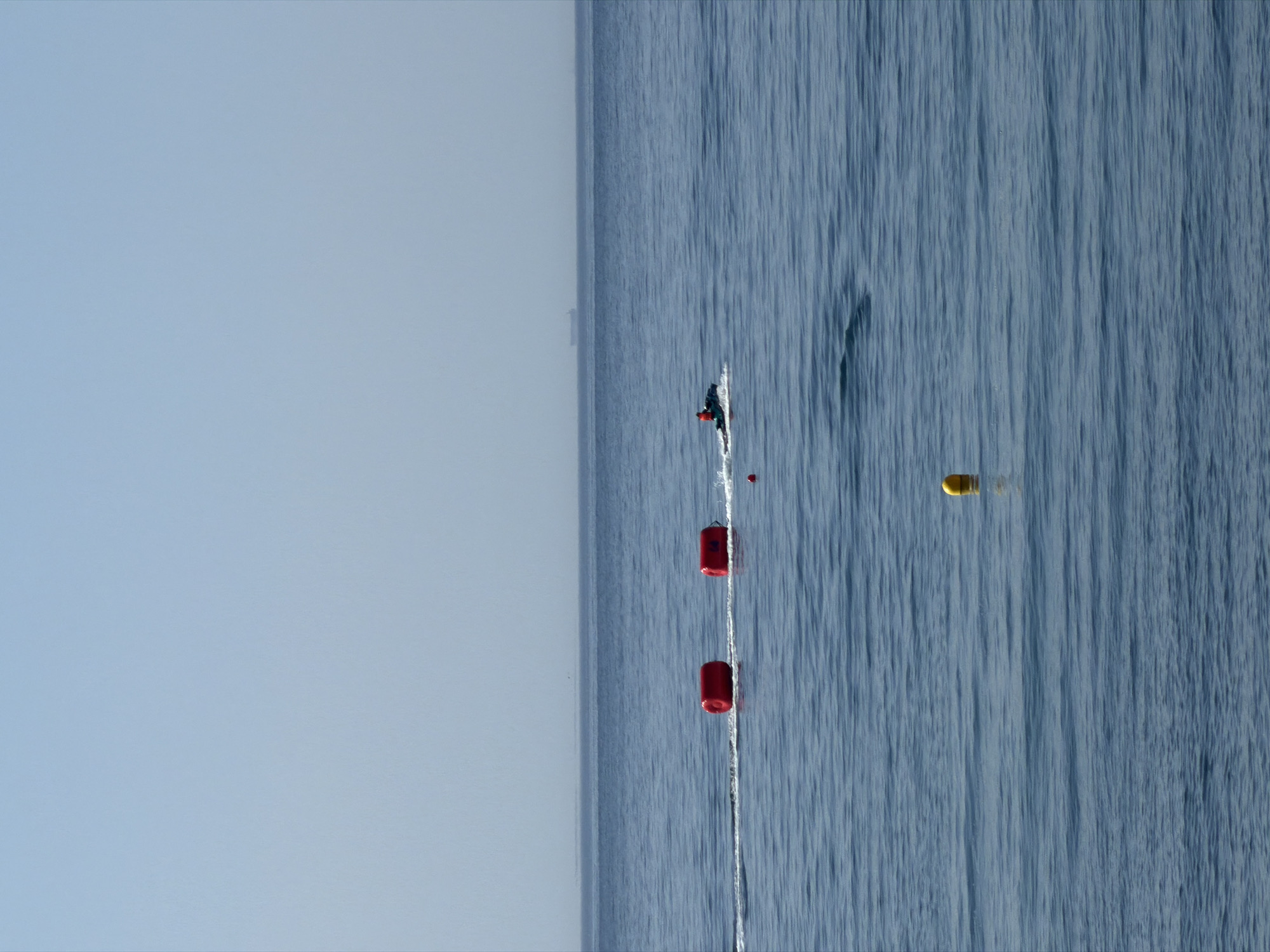 This image captures a serene ocean scene off the coast of Estepona, Spain, under a clear, pale blue sky. The calm, deep blue water stretches to the horizon, where a distant cargo ship is faintly visible. In the mid-ground, two cylindrical red buoys float side-by-side, leaving a wake on the water. To the right of the buoys, a person on a jet ski is actively moving through the water, also creating a trail. A smaller red buoy is positioned closer to the jet ski. Further to the right, a lone yellow buoy bobs gently on the surface. The overall impression is one of open water with recreational and navigational markers.