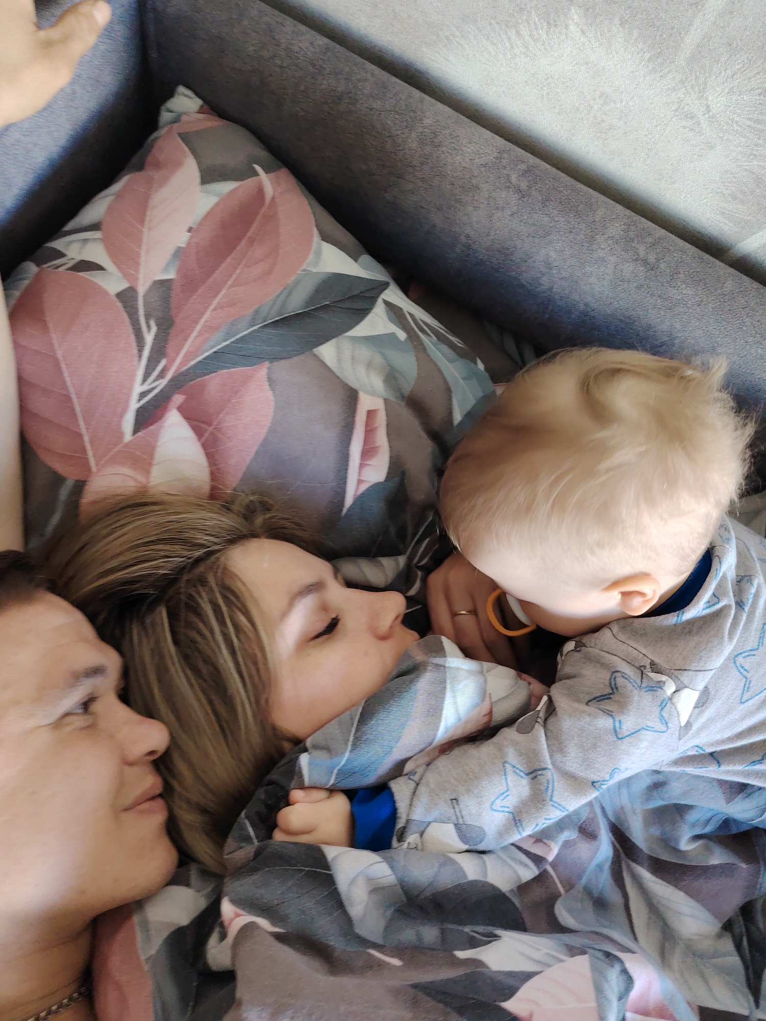 This image appears to have been taken in a private residence, likely in the city of Chaikovsky, Russia. It shows a mother, father, and young child lying together on a bed or couch. The mother is lying with her head on a patterned pillow, and the father is lying beside her. The child is lying between them, with their face close to the mother's. The child is wearing a grey pajama set with blue stars. The scene suggests a moment of familial closeness and rest.
