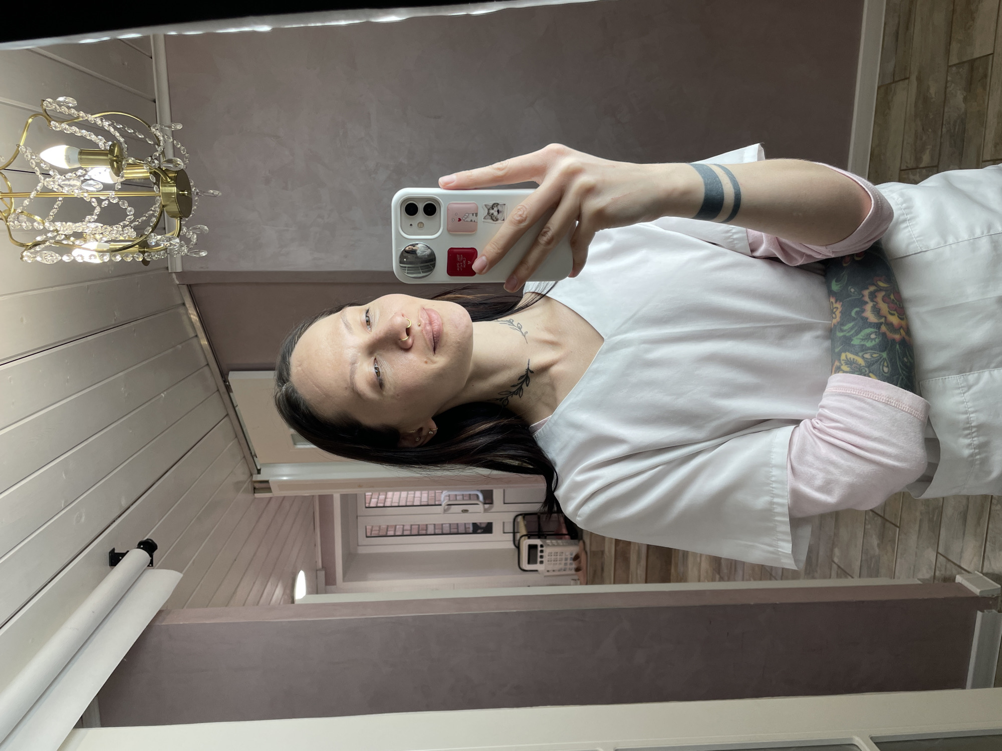 A woman is taking a selfie in a room with textured grey walls and white trim. She is wearing a white lab coat over a pink long-sleeved shirt. Her left arm has a tattoo of two parallel black bands, and her right arm has a floral tattoo. The phone she is holding is white with stickers on the back. A chandelier is visible in the top left corner. A hallway with windows and a phone is visible in the background. It is unknown if the location is Izhevsk, Russia.