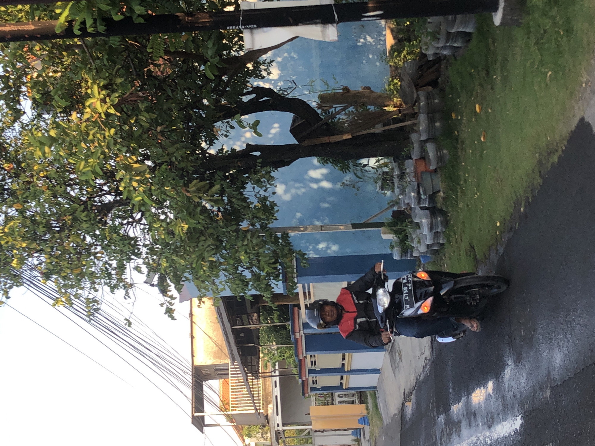 A person wearing a black and red jacket and a black helmet is riding a black motorcycle with a license plate reading "W 4798 RJ" and "06-23" in Kalidawir, Indonesia. The motorcycle has its headlights on and the rear turn signals are illuminated. The rider is wearing blue jeans and flip-flops. The motorcycle is on a wet asphalt road. To the right of the road is a grassy area with potted plants and some fallen leaves. Behind the grass is a blue wall with trees casting shadows. In the background, there is a building with an elevated porch and a light-colored roof, as well as power lines. The scene appears to be a street in a residential area during daylight.