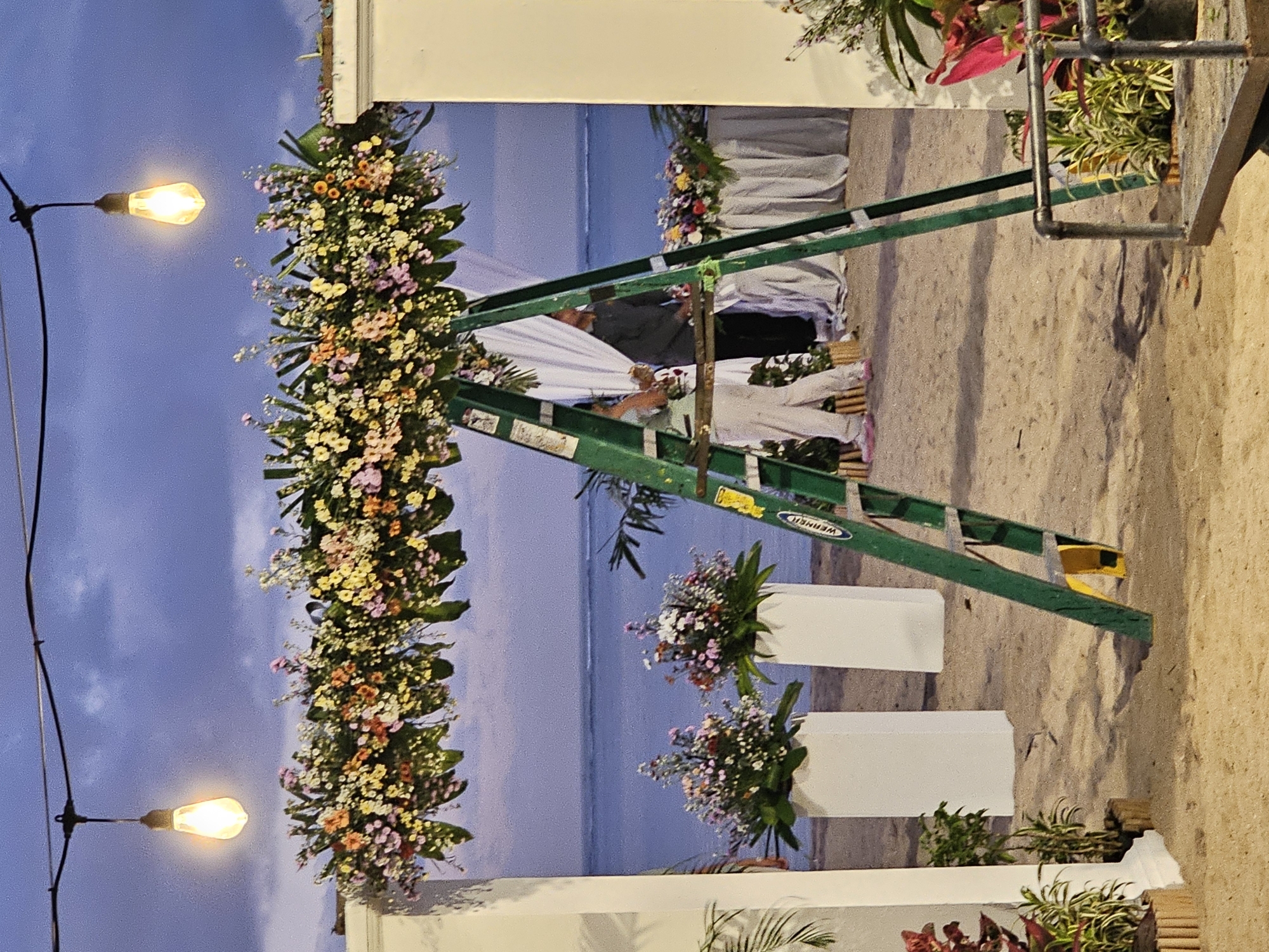 The image shows an outdoor wedding setup at what appears to be a beach location. The sky is dusk-colored. The wedding archway is decorated with dense floral arrangements in colors of white, yellow, orange, pink, and green. To the right, a green ladder is leaning against the arch structure. White rectangular planters with floral arrangements are also present. In the background, people are seated on chairs. String lights hang from the left side. The location may be Denpasar, Indonesia.