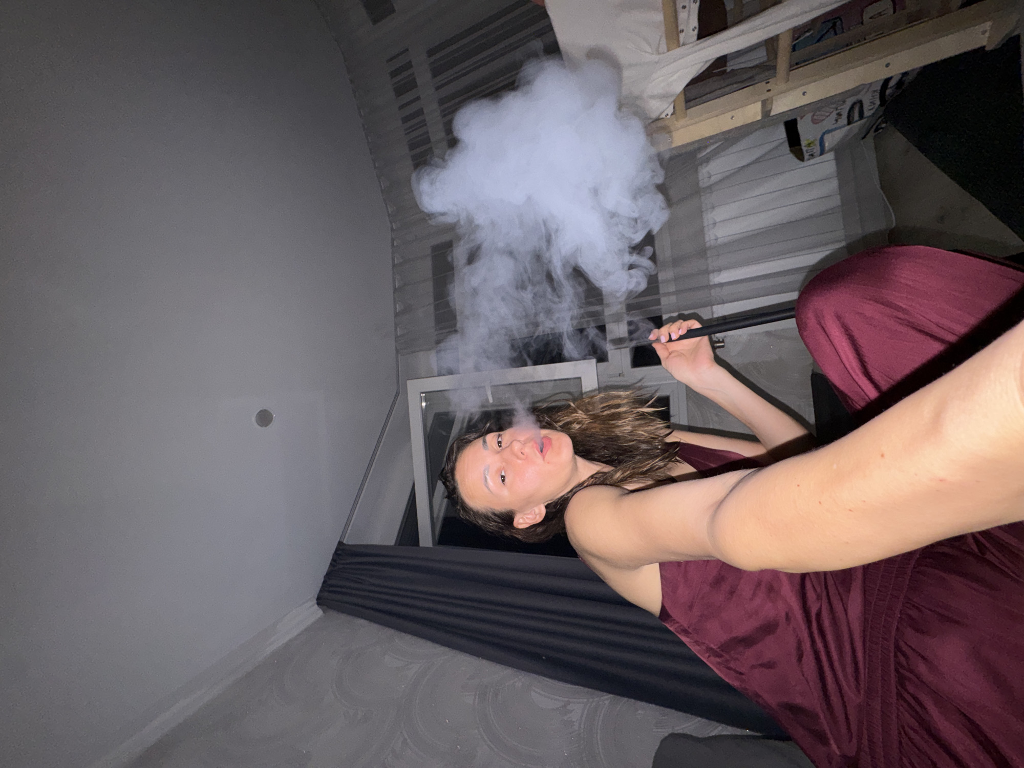 A low-angle, selfie-style photograph captures a young adult female indoors in Sochi, Russia, at night or late evening. She has medium-length brown hair, appears to have white residue on her forehead and eyebrows, possibly from a facial treatment. She is wearing a dark red/burgundy sleeveless top or dress. Her right arm is extended towards the camera, and she is holding a black hose or mouthpiece, presumably from a hookah or vape device, from which she is exhaling a large, dense cloud of white smoke to her right. The background features light gray walls, with a textured, wave-patterned wallpaper on the left. Dark gray curtains are drawn over a window on the left. A white-framed window is visible behind her, partially open to the dark exterior, with sheer white curtains covering another window to the right. A light-colored wooden frame, possibly part of a bunk bed or storage unit, is visible on the far right with white fabric draped over it. The ceiling is light gray and includes a single recessed light fixture.