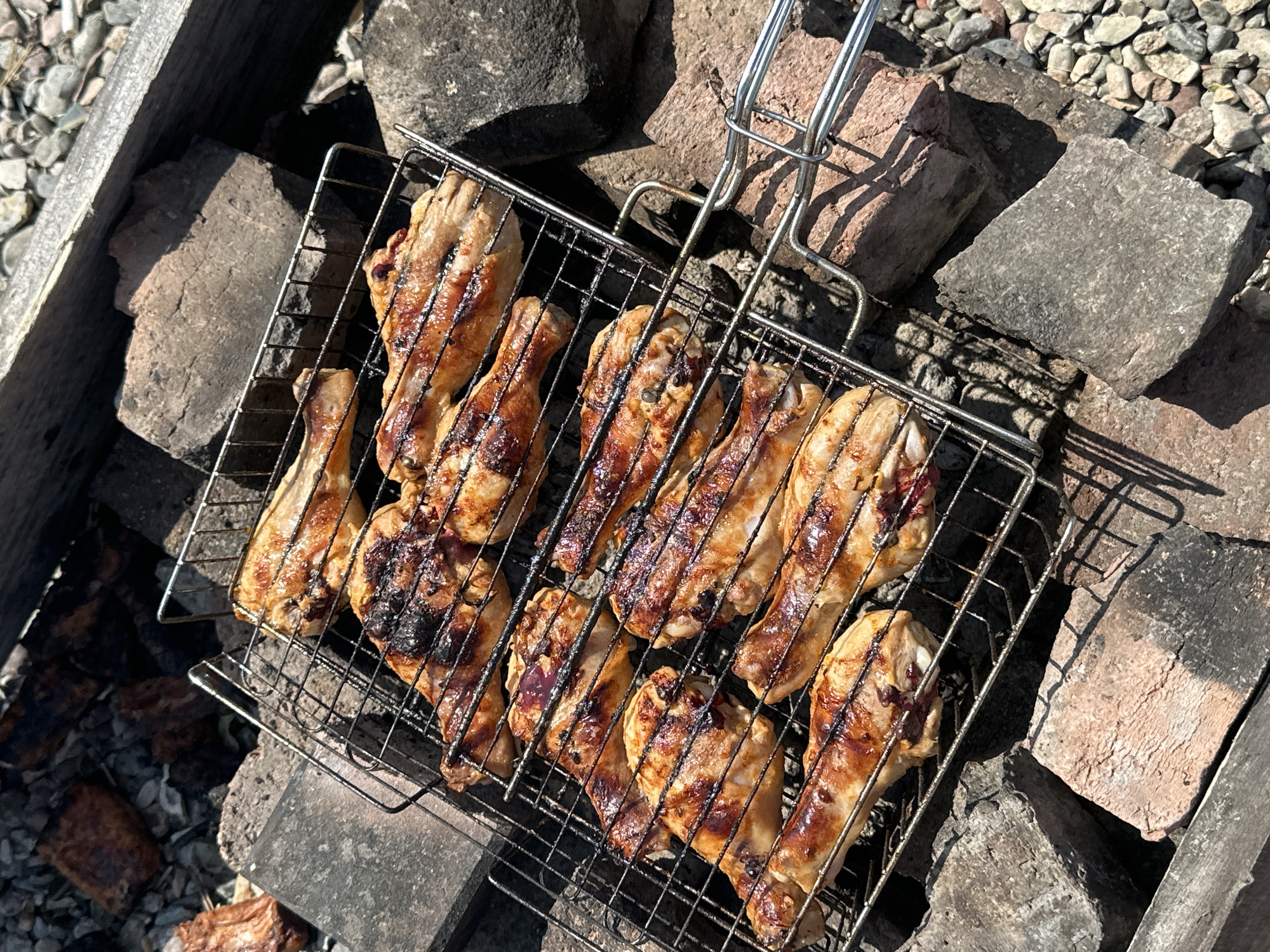 This is a top-down, close-up shot of several cooked chicken drumsticks on a metal grill basket. The chicken pieces are arranged in two rows within the basket and appear to be seasoned and grilled to a golden brown with some areas showing charring. The grill basket has a hinged handle, suggesting it is portable. The basket is placed on a rough, uneven surface composed of large stones and what appear to be bricks, creating a rustic outdoor setting. In the background, there are smaller stones and gravel. The lighting suggests it is daytime, with distinct shadows cast by the grill and chicken, indicating a sunny environment. The scene is likely taking place outdoors, possibly at a campsite or a backyard barbecue. No people are visible, and there is no visible text in the image. The overall impression is one of a prepared meal ready to be served.