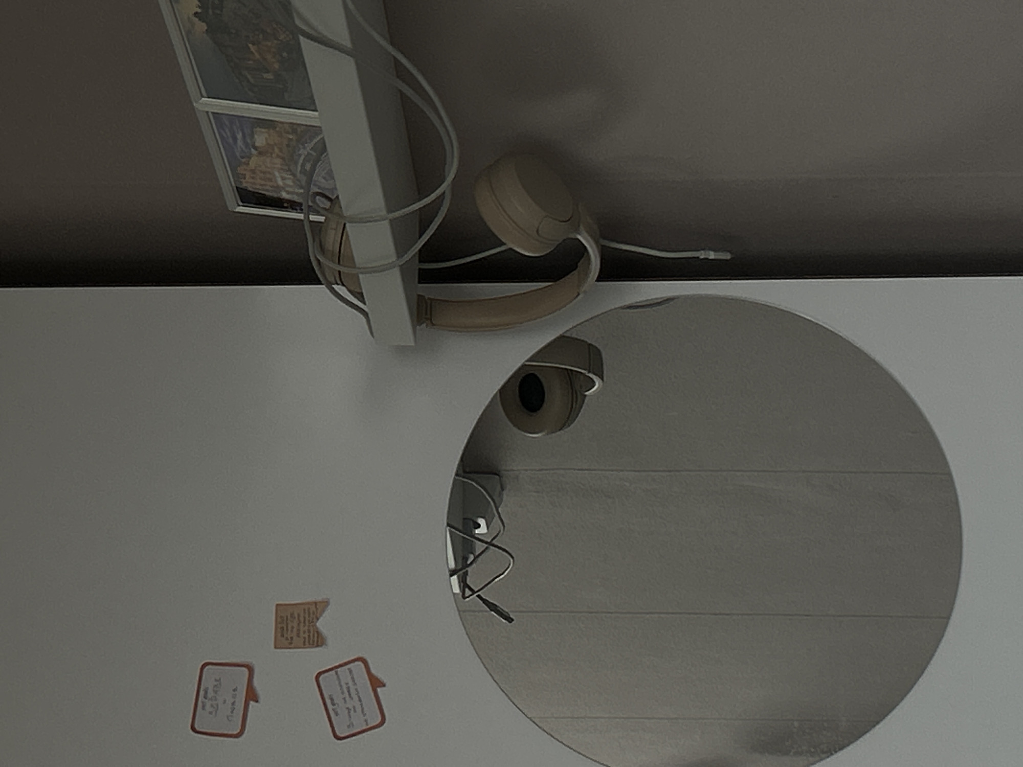 The image shows a white surface with several items. A pair of beige headphones is located on the upper right. Adjacent to the headphones is a picture frame. Below the headphones is a round mirror, reflecting another set of headphones and a cable. Three small note papers are affixed to the white surface in the lower-left corner. The image does not provide enough information to determine the location in Sofia, Bulgaria.