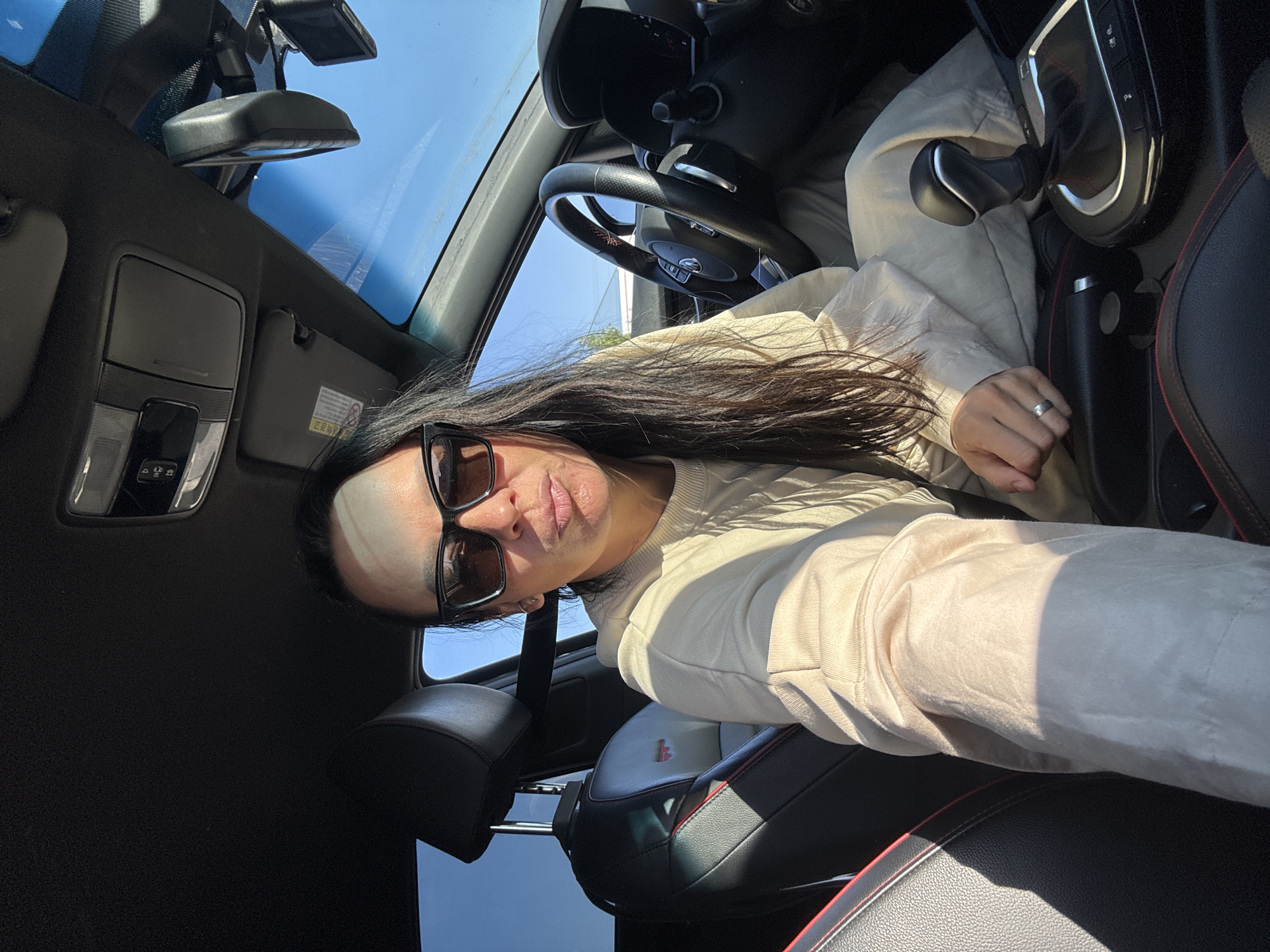 A person with long dark hair and sunglasses is taking a selfie from inside a car. The car has black leather seats with red stitching, a black steering wheel, and a black gear shift. The sunroof is open, and a device is mounted near the rearview mirror. The location in Chaikovsky, Russia cannot be determined from this image.