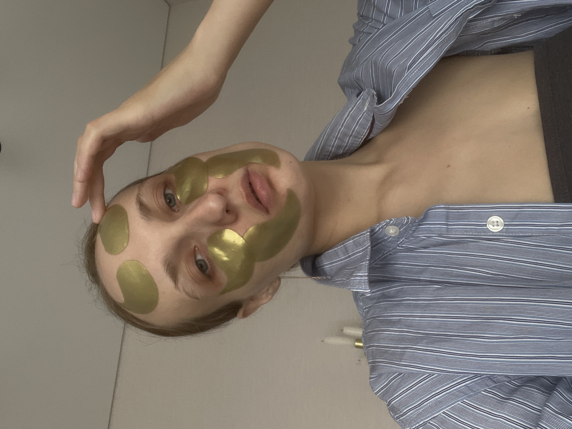A woman is lying on her side, wearing a blue and white striped shirt and gold eye and face masks. Her left hand is raised above her head. The background consists of white walls. The location is not discernible as Подшивалово, Russia based on the image content.