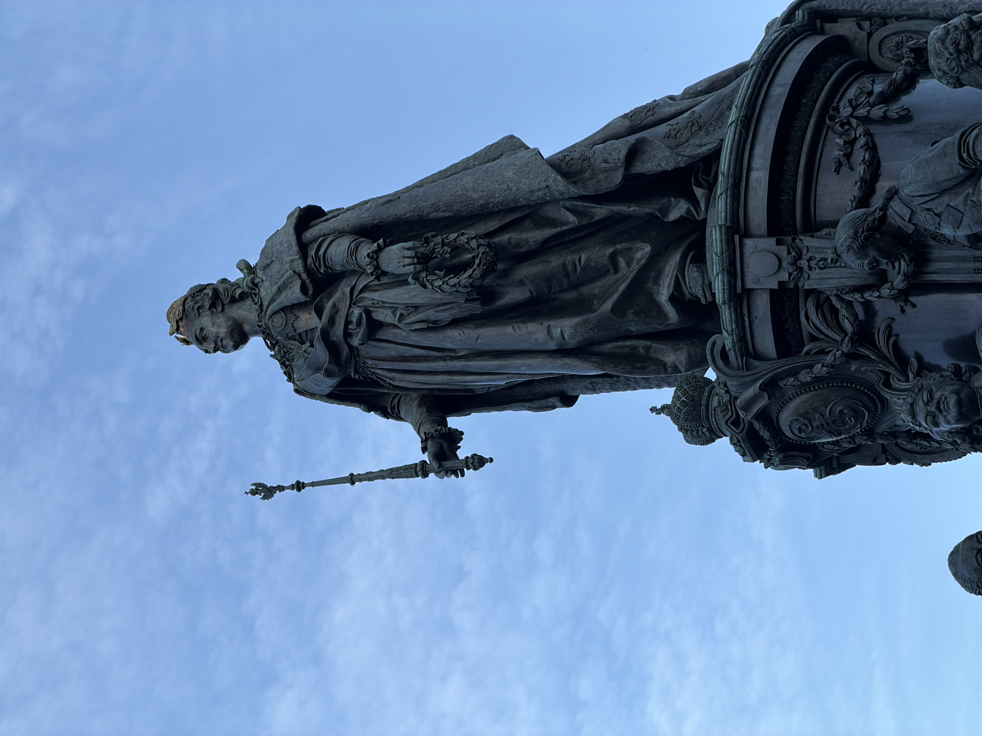 The image shows a low-angle view of the Monument to Catherine the Great in Saint Petersburg, Russia. The bronze statue of Catherine the Great stands tall, holding a scepter in her right hand and a laurel wreath in her left. She is adorned with a crown, a cape, and elaborate clothing. The base of the monument is decorated with relief sculptures of prominent figures from her reign. The monument is set against a backdrop of a cloudy blue sky.