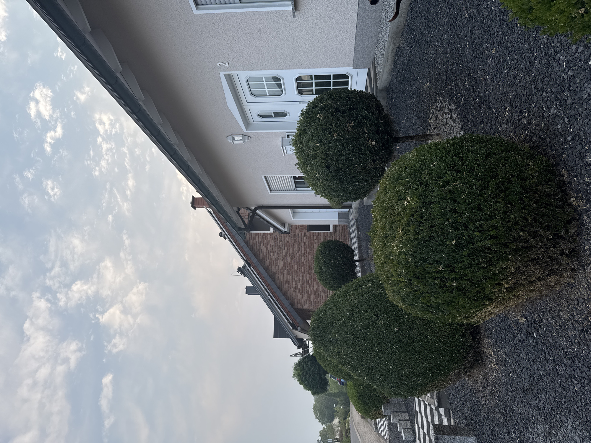 The image shows a row of houses in Überherrn, Germany, under a cloudy sky. The foreground is dominated by meticulously pruned, dark green bushes and dark grey gravel. To the right, a light beige house has a white double door with a glass pane and the number "2" above it. A white outdoor lamp is mounted to the left of the door. Next to this house, a house with a brick facade and a dark roof is visible. Further down the street, more houses and trees can be seen, with a road on the left. The overall impression is one of a well-maintained residential area.