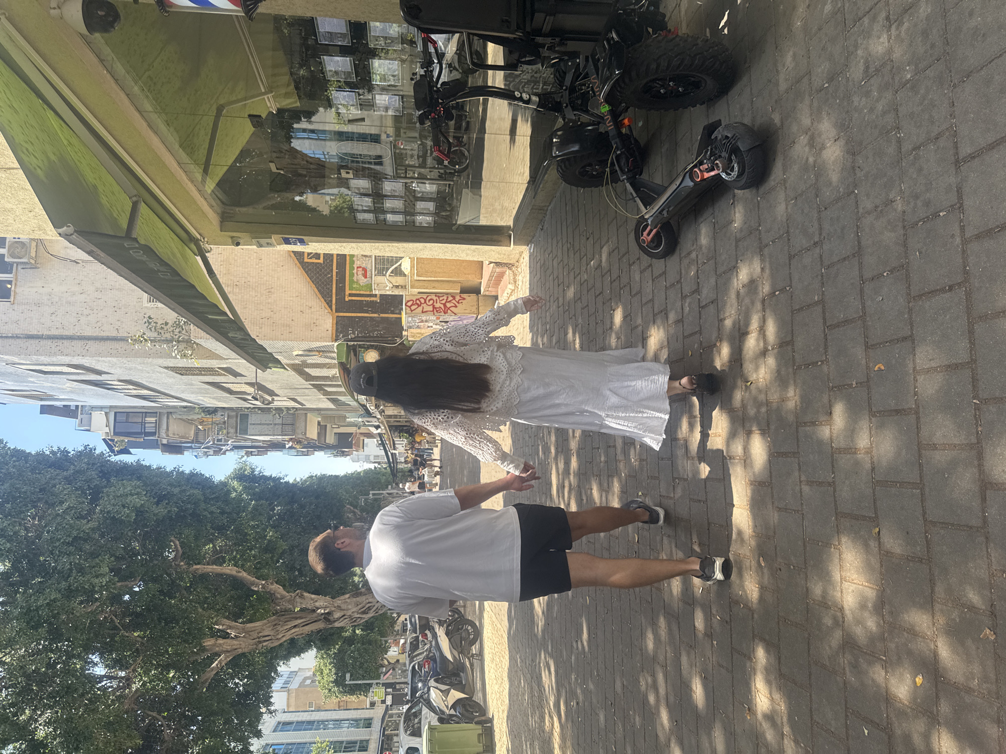 A side-view shows a couple walking hand-in-hand on a brick-paved sidewalk in a city. The woman is in a long white dress, and the man is wearing a white t-shirt and black shorts. A scooter is parked near the couple. Nearby trees create shadows on the sidewalk. Buildings are visible on either side of the street. The image likely depicts a street in Tel-Aviv, Israel.