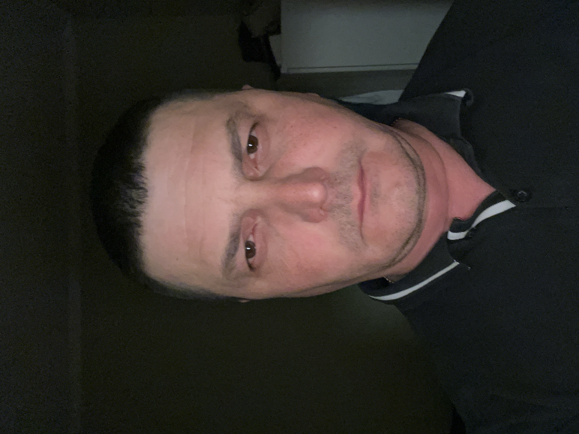 A close-up, indoor photograph shows a middle-aged man with short, dark hair and light skin. He is wearing a dark polo shirt with a white stripe at the collar. The location is unknown.