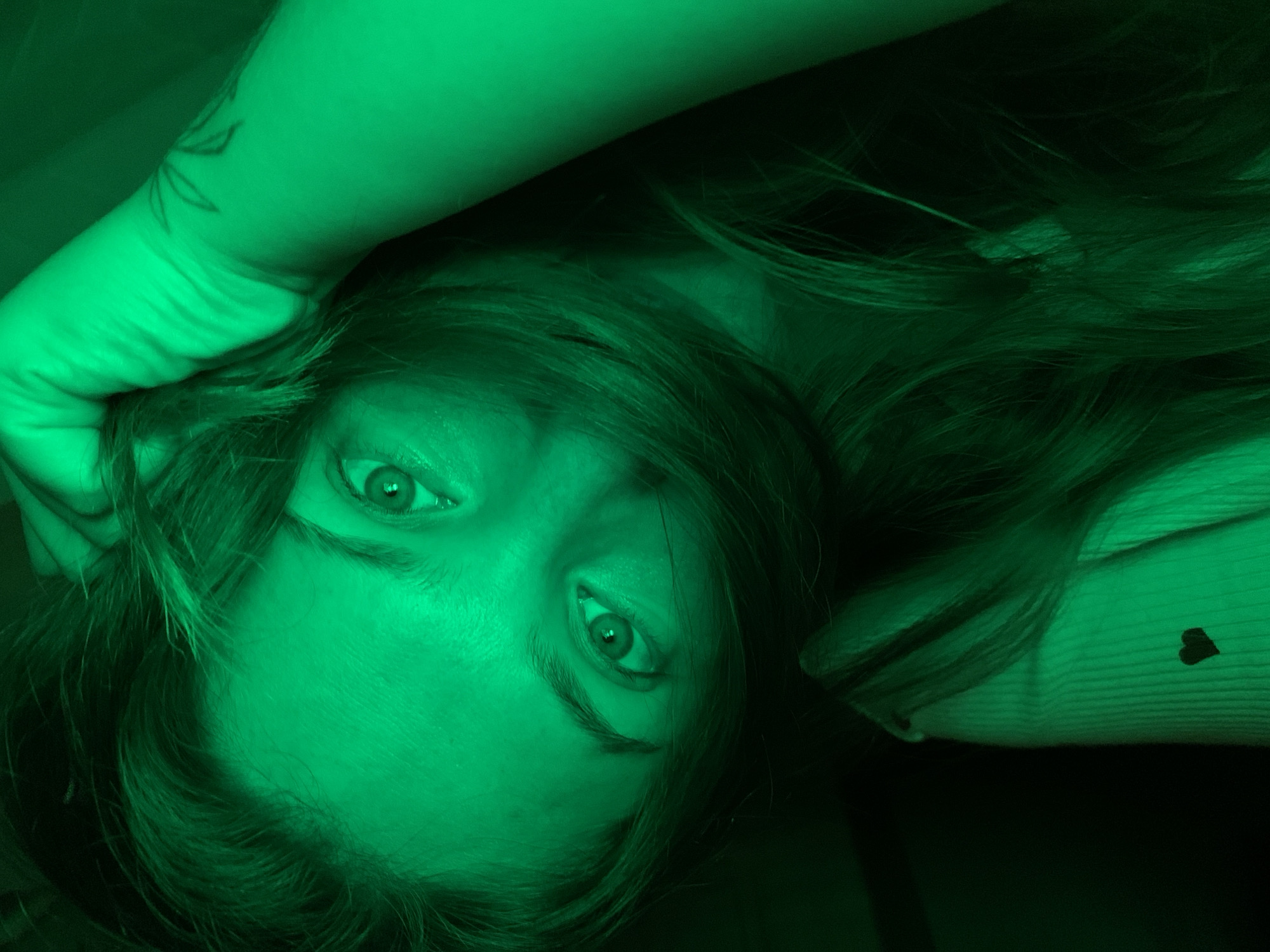 A low-angle, close-up, indoor selfie of a young woman with green lighting. She is looking directly at the camera, with her right hand near her head, partially obscuring her face with her hair. She is wearing a light-colored, ribbed shirt with a small dark heart design on the left side of her chest. The background is dark and out of focus. There is a tattoo visible on her forearm. The scene is likely a private residence in Chaikovsky, Russia.