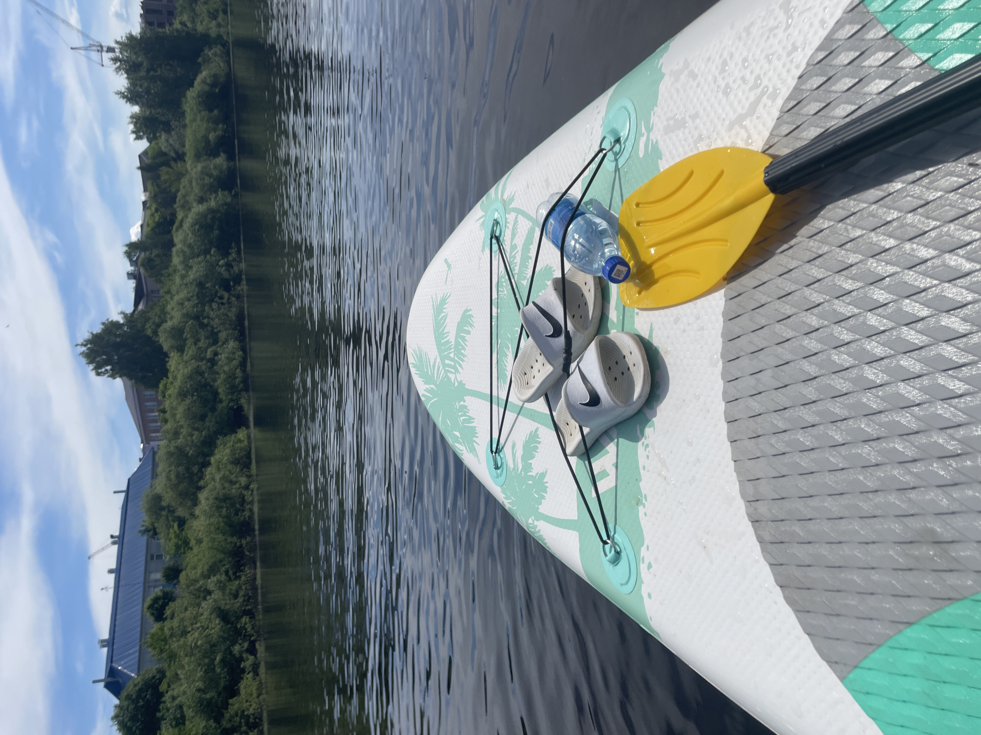 The scene depicts a forward-facing view from a stand-up paddleboard (SUP) on the Vologda River in Vologda, Russia, under a clear, partially cloudy sky.
In the foreground, the white deck of a SUP occupies the lower portion of the image. It features a gray, diamond-patterned textured grip pad in the center, flanked by white and teal sections with a repetitive teal palm tree graphic. Black bungee cords are stretched across the front deck, secured by teal D-rings. Placed beneath these cords are two white Nike slide sandals with black swoosh logos. To their right, a translucent blue plastic water bottle with a white cap and a square QR code sticker is also secured. A yellow paddle blade, attached to a black shaft segment, rests on the gray grip pad to the right of the water bottle.
The midground is dominated by the calm, dark surface of the river, exhibiting subtle ripples that reflect the overhead sky. The background reveals a distant riverbank densely covered with lush green deciduous trees and bushes. Above the treeline, several structures are visible: a long building with a distinctive dark blue corrugated metal roof and light-colored walls is prominent to the left. Further to the right, a large construction crane with an extended jib rises above the trees and a reddish-brown building, indicating ongoing development. The sky above is bright blue with scattered white cumulus clouds. No individuals are visible in the frame.