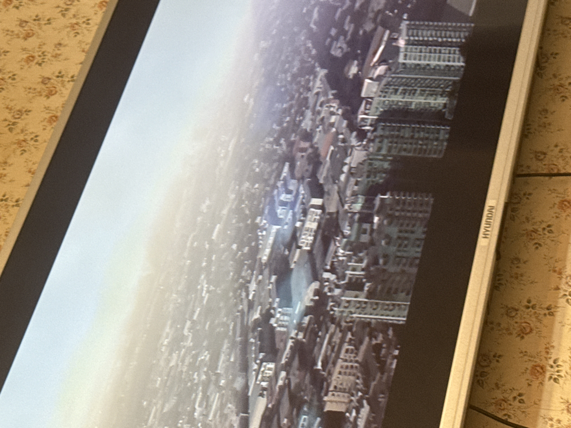 The image depicts an aerial view of a city, with numerous buildings and structures visible. The perspective is tilted. The city appears to be densely populated. The TV's bezel and Hyundai logo are visible. The background behind the TV features a floral-patterned wallpaper. Location in the city of Mytishchi, Russia is impossible to determine from the image.