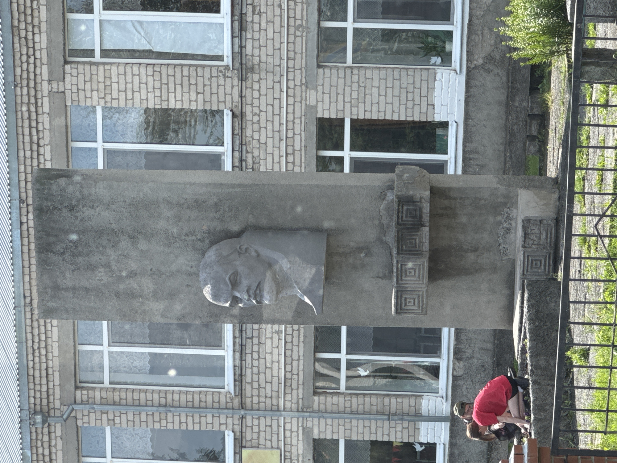 The image depicts a bust of Vladimir Lenin mounted on a grey concrete pillar in front of a brick building. The building has several white-framed windows. In the foreground, two young individuals, one wearing a red shirt and black shorts, and another wearing a green cap and dark clothing, are interacting near the base of the pillar, appearing to be engaged in some activity on the ground. A black metal fence runs across the bottom of the frame, with grass and pavement visible behind it. The scene is set outdoors, likely in a public area, given the presence of the monument and the building.