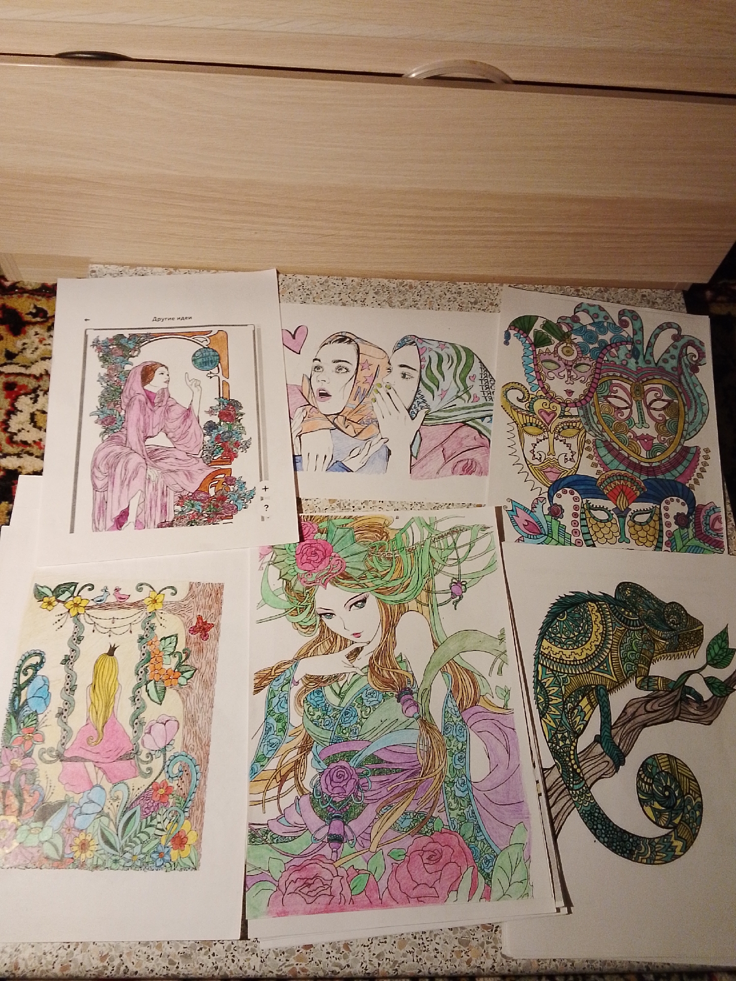 The image shows a collection of six colored drawings on white paper, arranged haphazardly on a surface that appears to be a tabletop or a counter, possibly made of a mottled stone-like material. The backdrop is a light-colored cabinet or storage unit, possibly made of wood, with a visible handle. The drawings depict various subjects, including a woman in a robe touching a globe, two women whispering, a stack of ornate masks, a woman with flowers, a girl on a swing surrounded by flowers, and a chameleon on a branch. The coloring style is amateur, with varying degrees of detail and completeness.
There is no indication of the location being the city of 1-Go Maya, Russia, in the image. There are no people visible in the frame, only the subjects of the drawings. The actions and interactions are limited to the content of the drawings themselves. The environment is a domestic setting, judging from the furniture.