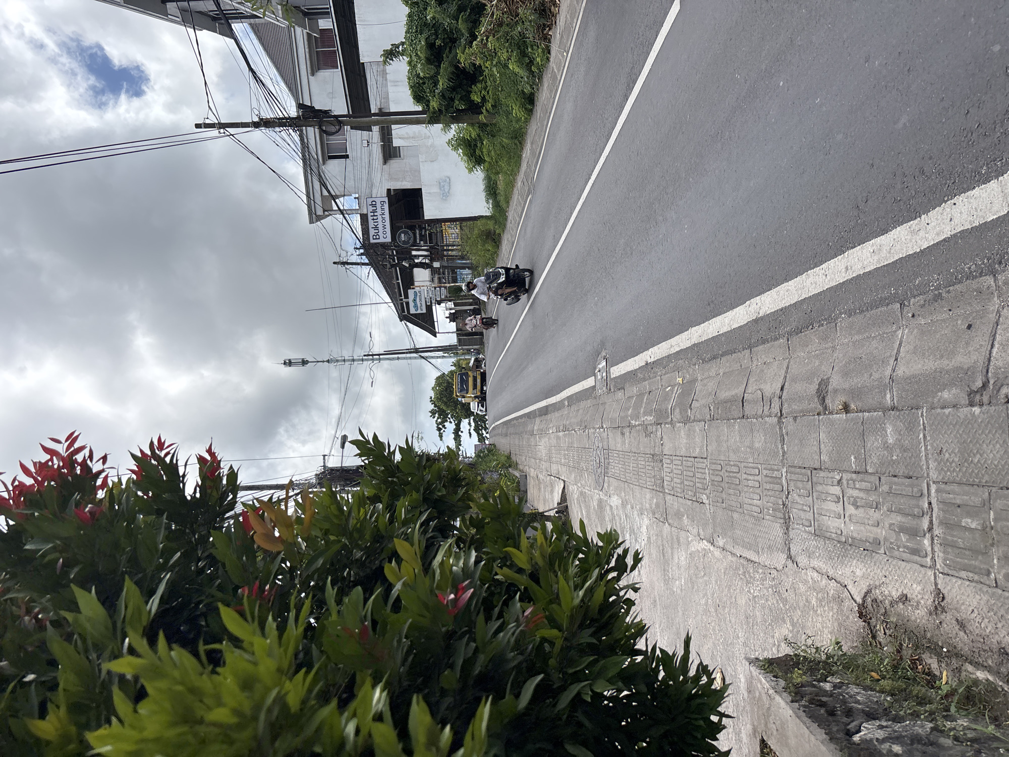 The image depicts a street scene in Kuta Selatan, Indonesia. A gray asphalt road with a white painted line along its edge stretches into the distance. A gray tiled sidewalk lines the road's edge with tactile paving blocks present. A lush bush with green and red leaves is visible in the foreground. On the left side of the road, a building with a sign for "BukitHub Coworking" is seen. Overhead power lines crisscross the sky, which is overcast with gray clouds. A yellow truck and a couple of motorcycles are visible on the road. Two people are on one of the motorcycles.