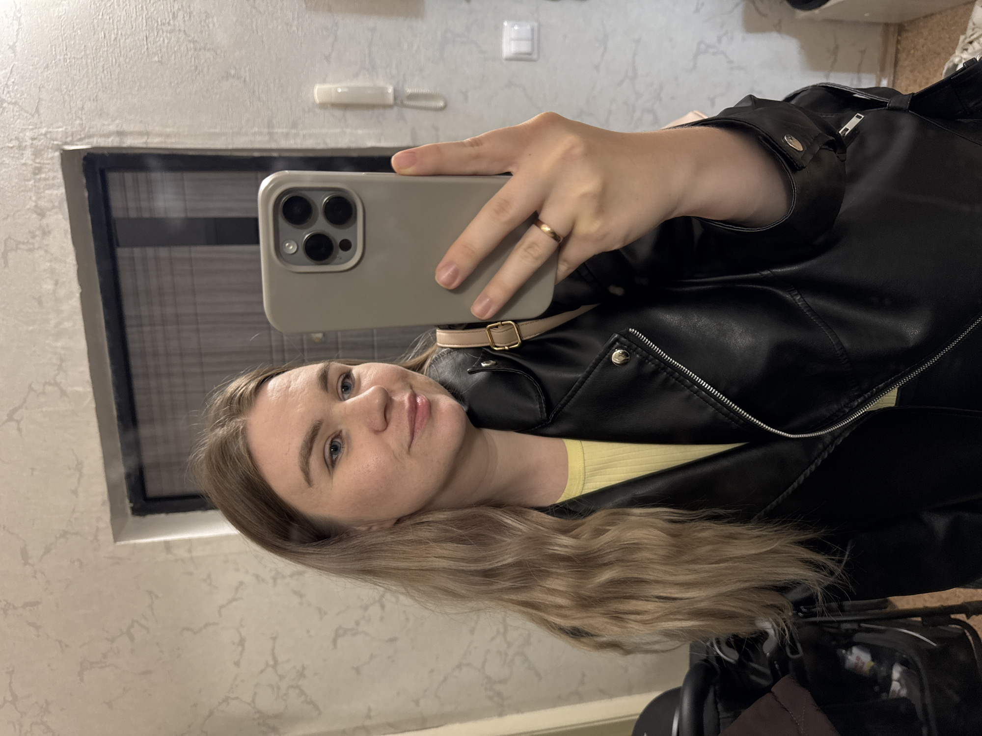 A woman is taking a selfie indoors, holding a cell phone in her right hand. She has long blonde hair and is wearing a black leather jacket over a yellow top. A gold ring is visible on her finger. The background includes a marble-patterned wall and a window with blinds. The setting appears to be a residential space. There is no information in the image indicating the city of Chaikovsky, Russia.