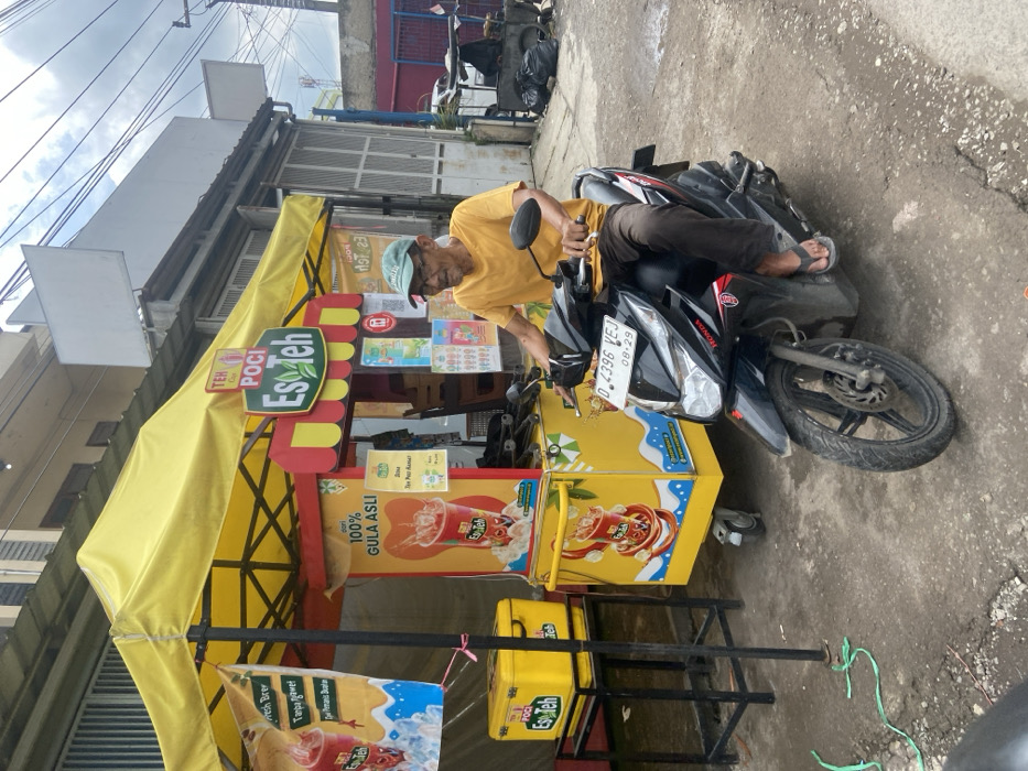 The scene takes place in Ciparay, Indonesia, at the coordinates -7.029 latitude and 107.698 longitude. An older man with a light complexion, wearing a yellow shirt, blue jeans, and a green hat, is sitting on a black Honda motorcycle with a red logo. He has a thin white beard and appears to be looking at the camera. The motorcycle is parked next to a bright yellow food cart. The cart has an awning that is a yellow and white striped pattern. The front of the cart has a red and yellow sign that says "Teh Poci Es Teh," and the sides have images of bottled drinks and the words "100% Gula Asli." The motorcycle has a license plate that says "D 4396 VEJ 08-29." The man is holding the motorcycle's handlebars with his right hand and his left hand is resting on his lap. The cart is facing a building to the right, which has a large white sign hanging above it. The building appears to be made of concrete and has a gray and black roof. There is another smaller building to the left of the food cart. The scene is outdoors, on a paved street, and there are two trash bags lying in the distance.