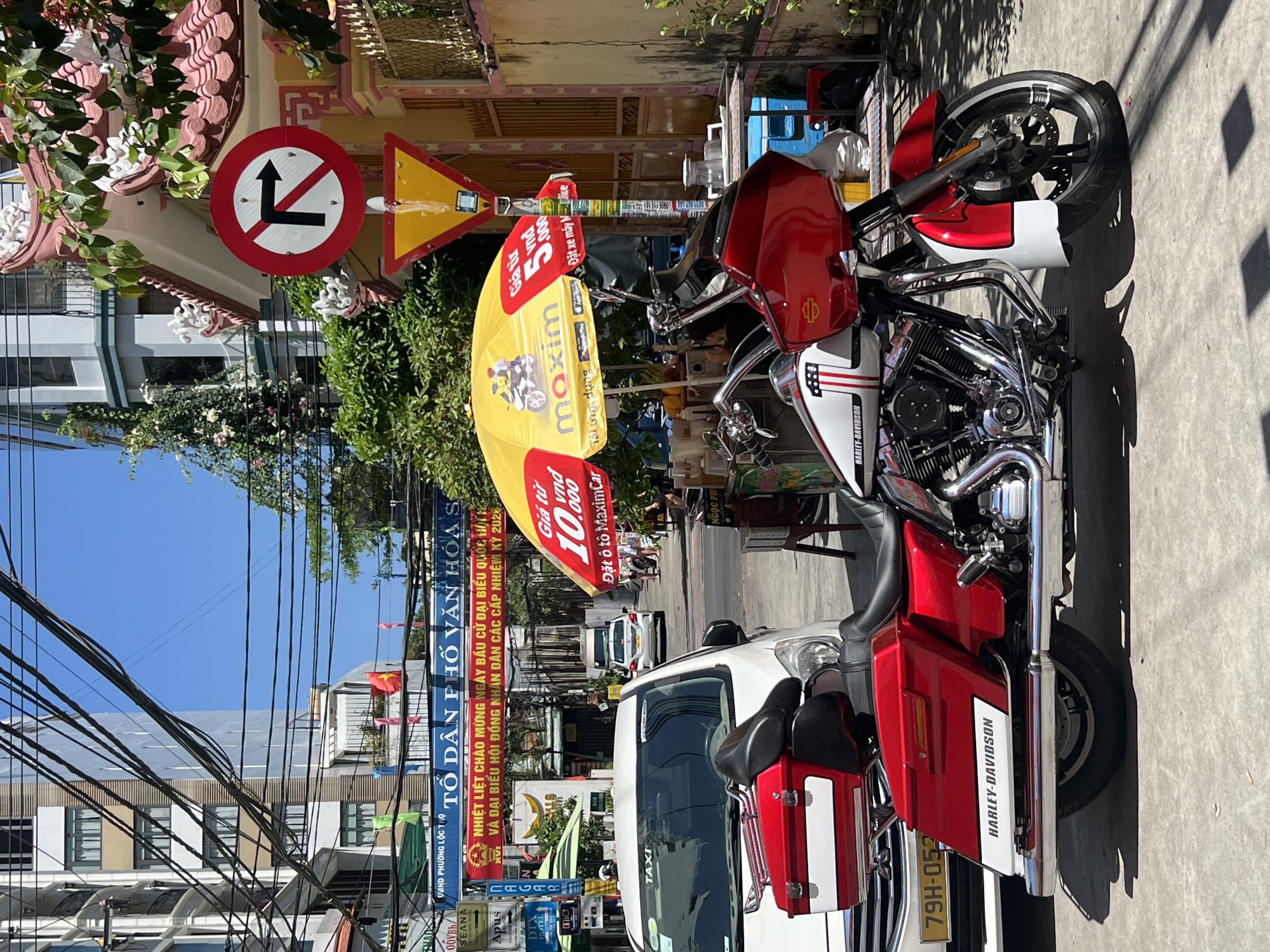 This image depicts a sunlit urban street scene in Nha Trang, Vietnam, under a clear blue sky.
In the immediate foreground, a red and white Harley-Davidson touring motorcycle is parked on a light grey paved surface, oriented towards the right. The motorcycle features a prominent fairing, saddlebags, chrome engine components, and "HARLEY-DAVIDSON" text on its white sections. Behind the motorcycle, the front portion of a white taxi vehicle, identified by "TAXI" on its windshield and a partially visible license plate starting with "79H-052", is parked.
To the right of the motorcycle, a large yellow umbrella, branded "Maxim", is open, providing shade. The umbrella advertises ride-sharing services, stating "Giá từ 10.000 vnd Đặt ô tô MaximCar" (Prices from 10,000 VND Order car MaximCar) and "Giá từ 5.000 vnd Đặt xe máy MaximBike" (Prices from 5,000 VND Order motorbike MaximBike). Beneath this umbrella, a street vendor setup includes a table with various containers and a blue cooler. The back of a seated individual is partially visible at this vendor station. Adjacent to the vendor, a red circular "No Right Turn" traffic sign is mounted on a pole above a yellow triangular "Yield" sign. These signs are positioned in front of a building characterized by a red-tiled, ornate roof and yellow exterior walls.
The street extends into the background, where additional vehicles, including a white van and a white car, are visible. Several pedestrians are present on the right sidewalk further down the street. The thoroughfare is flanked by multi-story buildings of varying architectural styles. A dense network of black electrical and communication cables is strung overhead against the clear blue sky. Two prominent red and white banners with Vietnamese text span across the street; one reads "TỔ DÂN PHỐ VĂN HÓA S" (Cultural Residential Group S) and another commemorates the election day for national assembly and people's council delegates for the 2021-2026 term. Green vegetation, including climbing plants and trees, is interspersed throughout the scene.