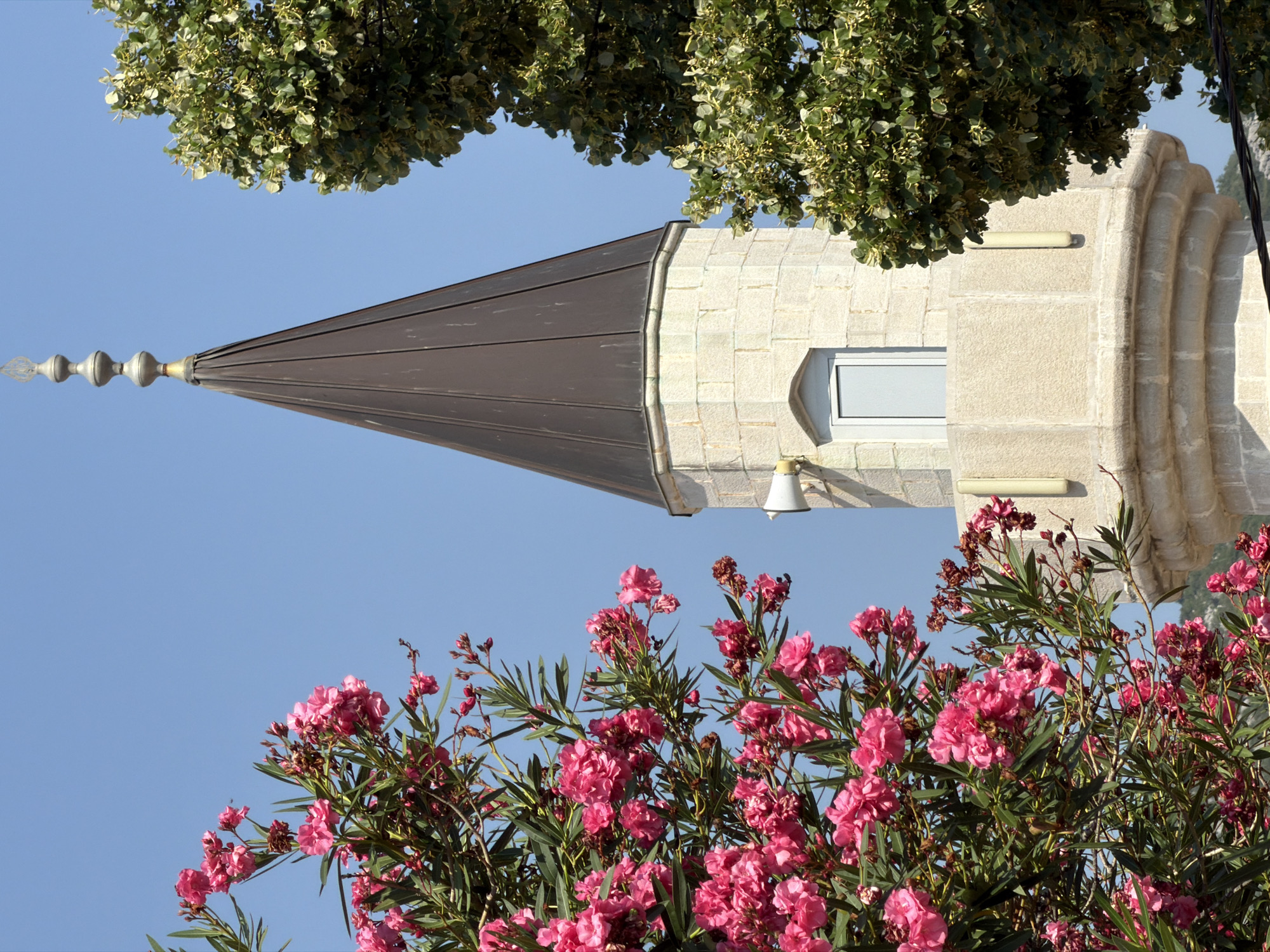 This is a close-up, eye-level outdoor shot of a minaret from a mosque in Trebinje, Bosnia and Herzegovina, framed by vibrant pink oleander flowers on the left and the lush green foliage of a deciduous tree on the right. The minaret is constructed of light-colored stone blocks with a textured finish. It has a rounded base and tapers upwards to a conical, dark brown metal roof topped with a finial. A small, light-colored rectangular window is visible on the minaret, and two light-colored loudspeakers are mounted on the exterior wall. The sky is a clear, bright blue. The focus is sharp on the minaret and the foreground flowers, with the tree foliage also relatively in focus. The overall impression is one of a sunny day with blooming vegetation.
