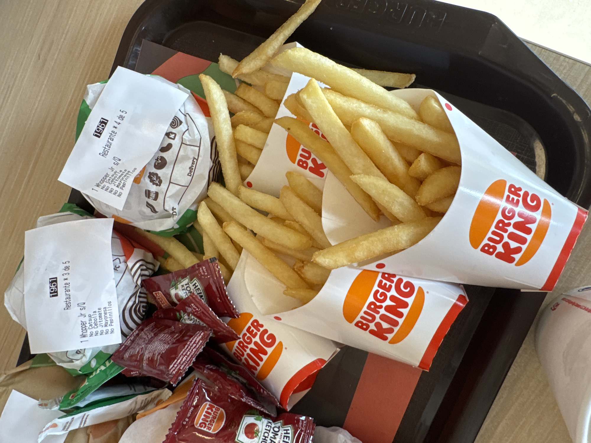 A top-down shot shows a black Burger King tray on a table. The tray contains three containers of Burger King french fries, a Burger King ketchup packet, a wrapper for a Whopper Jr., and two receipts from Restaurant 1961, indicating the location in the city is Barrio de Nuevo León, Mexico. The receipts indicate one order contains a Whopper Jr. with no ketchup, onion, tomato, mayonnaise, or pickles. The other order also contains a Whopper Jr., with no onion, tomato, mayonnaise, or pickles.