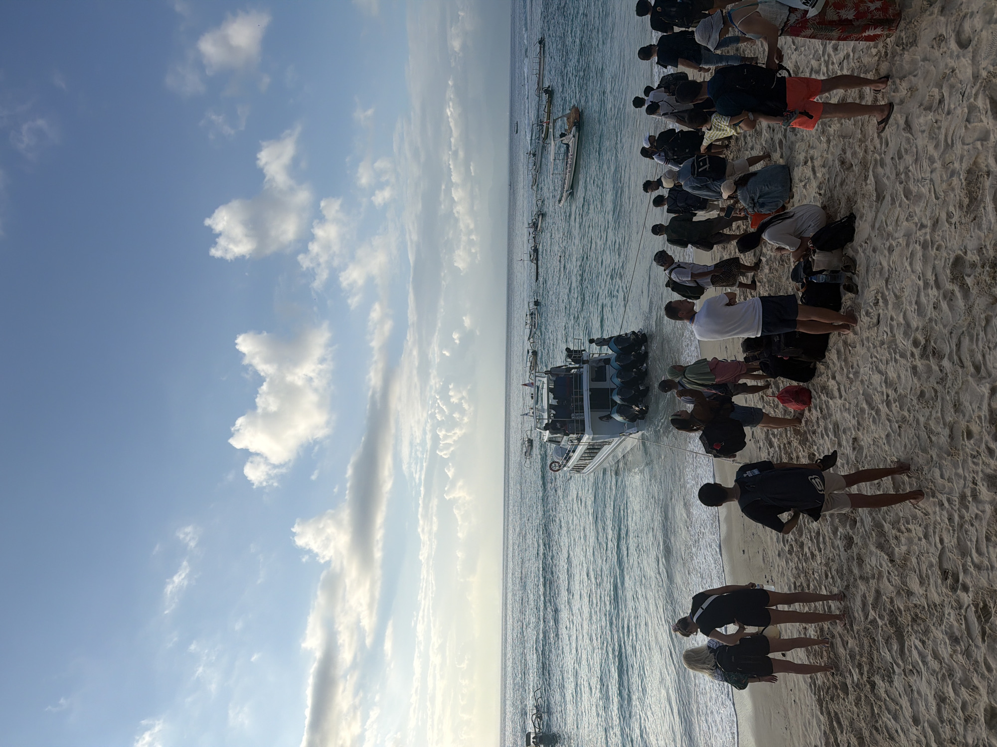 A group of tourists are gathered on a sandy beach in Nusa Penida, Indonesia, awaiting transport on a boat. The scene depicts a tropical coastline with calm turquoise waters under a partly cloudy blue sky. A white speed boat with multiple outboard motors is docked near the shore, and several other smaller boats are visible further out at sea. The people are diverse in age and gender, wearing casual summer attire. Some are carrying backpacks and luggage, indicating travel. The atmosphere appears relaxed, suggesting a transition point for travelers. The lighting indicates either early morning or late afternoon.