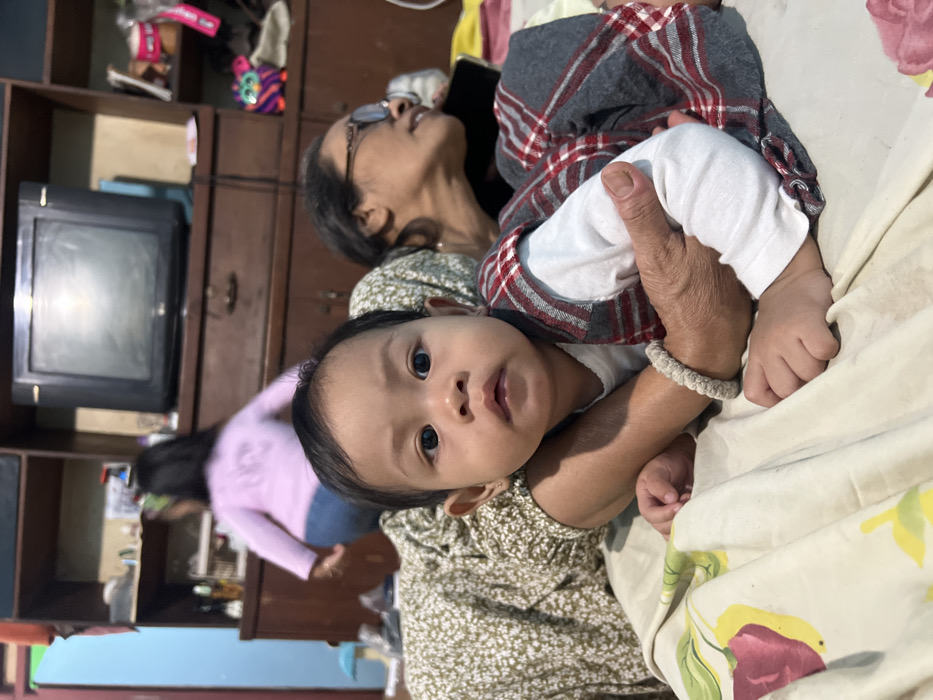 An indoor scene in a residential dwelling in Kecamatan Pati, Indonesia, features three individuals and a partially visible fourth. In the central foreground, an infant is positioned lying down, gazing directly forward at the camera. The infant wears a white long-sleeved shirt, a plaid grey and red sleeveless romper, and a light-colored braided bracelet on the right wrist. The infant's head is slightly elevated. A visible arm, belonging to an individual dressed in a light green garment with a white floral pattern, supports the infant. To the right of the infant, the head and shoulders of an adult female are visible, wearing glasses and looking towards the right side of the frame, away from the camera. In the background, further back and blurred, another child is standing or moving, facing away from the camera, wearing a light pink long-sleeved shirt and blue jeans. The subjects are positioned on a soft surface covered by a light-colored sheet with a floral pattern featuring red and yellow elements. Behind them, a dark brown wooden cabinet or shelving unit occupies the background, topped with an older model black cathode ray tube television. Various objects, including bottles, containers, and a colorful striped toy, are arranged on the cabinet shelves. A light blue mushroom-shaped object is visible on a lower surface to the left. A light blue wall is visible in the upper left background.